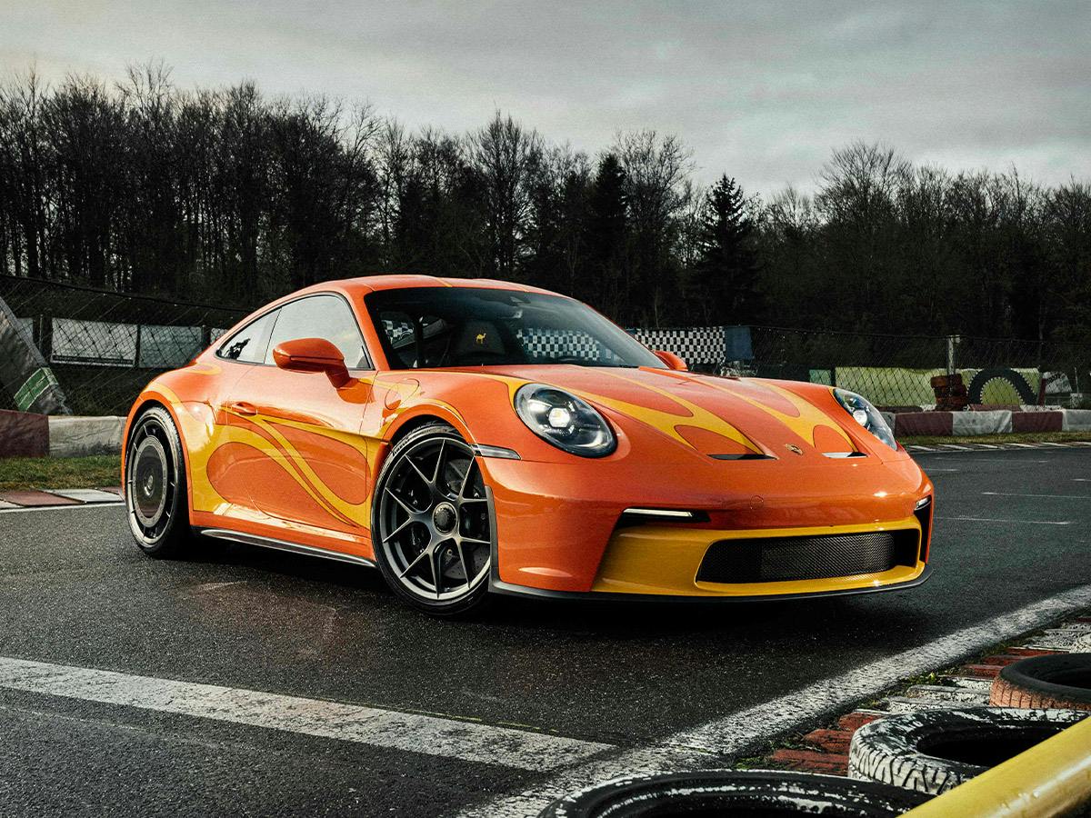 Porsche 911 S/T in Phoenixred with Signalyellow accents, photographed from a low front-side angle on a race track with tire barriers and a cloudy sky in the background.