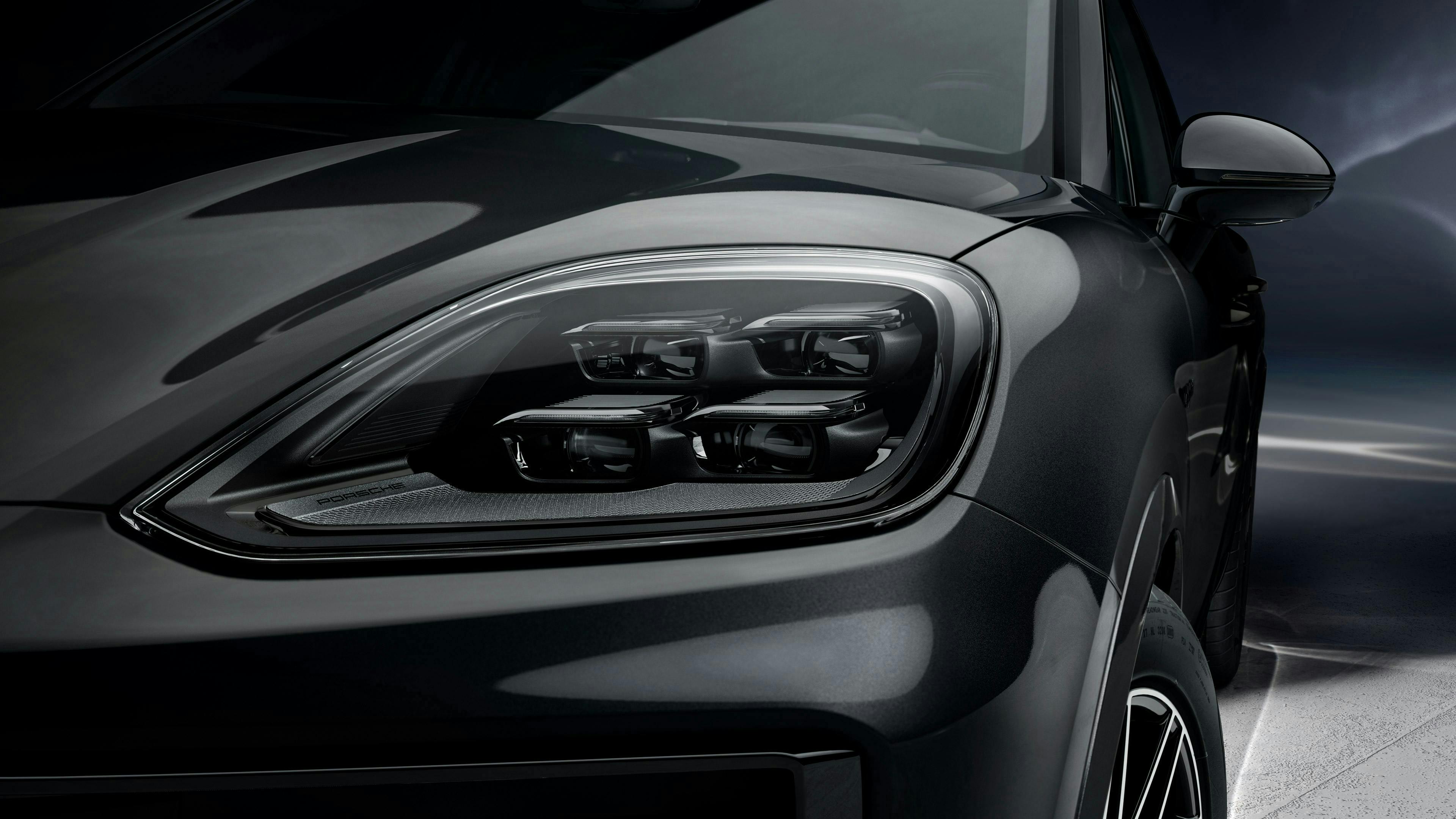 Czarne Porsche Cayenne Black Edition, widok z przodu, włączone reflektory LED, tło studyjne. Szare Porsche Cayenne, widok z tyłu, czarne felgi, zaparkowane przed wysoką ciemną ścianą. Zbliżenie lewego reflektora LED czarnego Porsche Cayenne, nowoczesny design świateł w studiu fotograficznym.