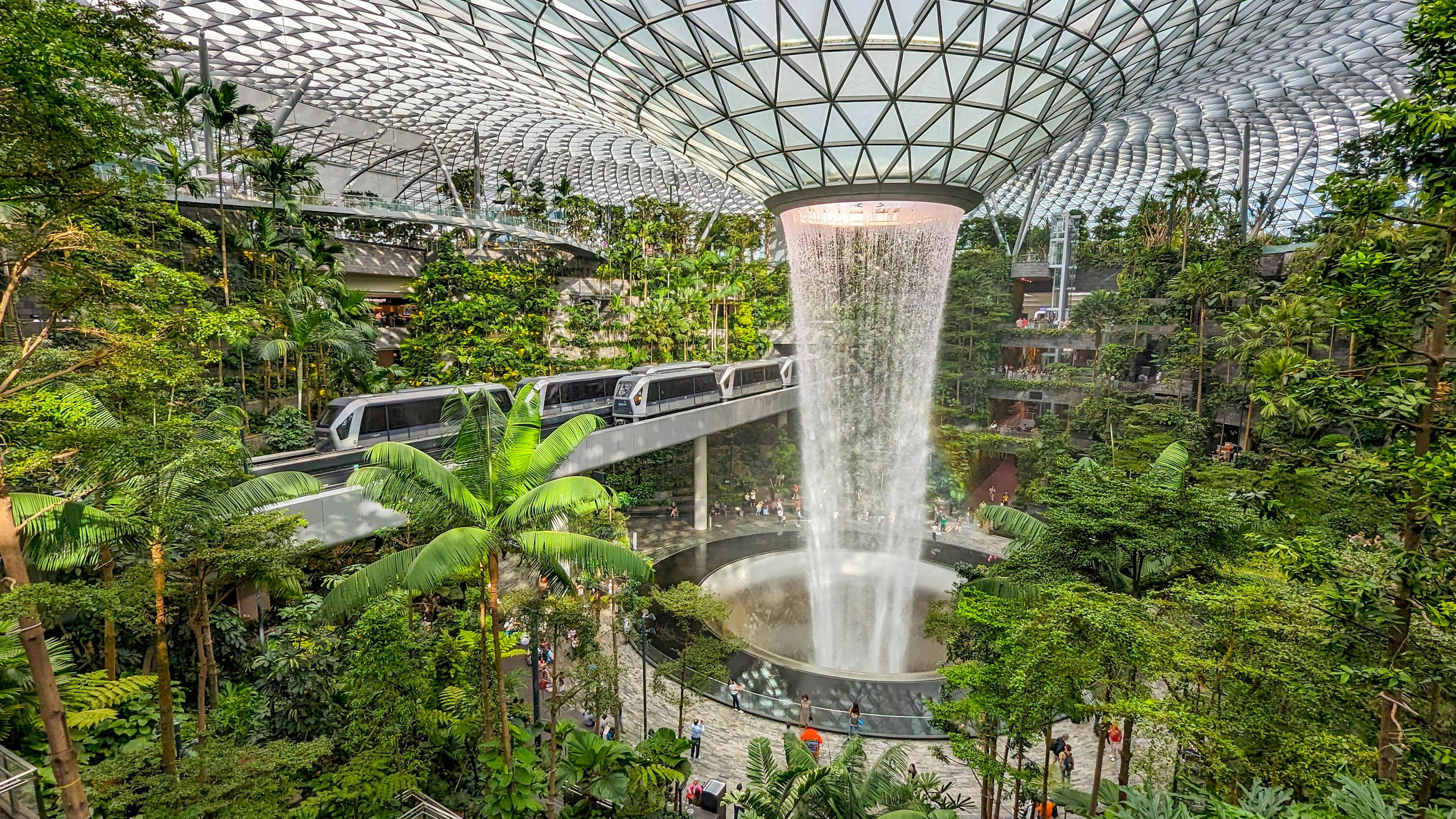 Indoor waterfall surrounded by lush greenery with a train passing through Jewel Changi Airport.