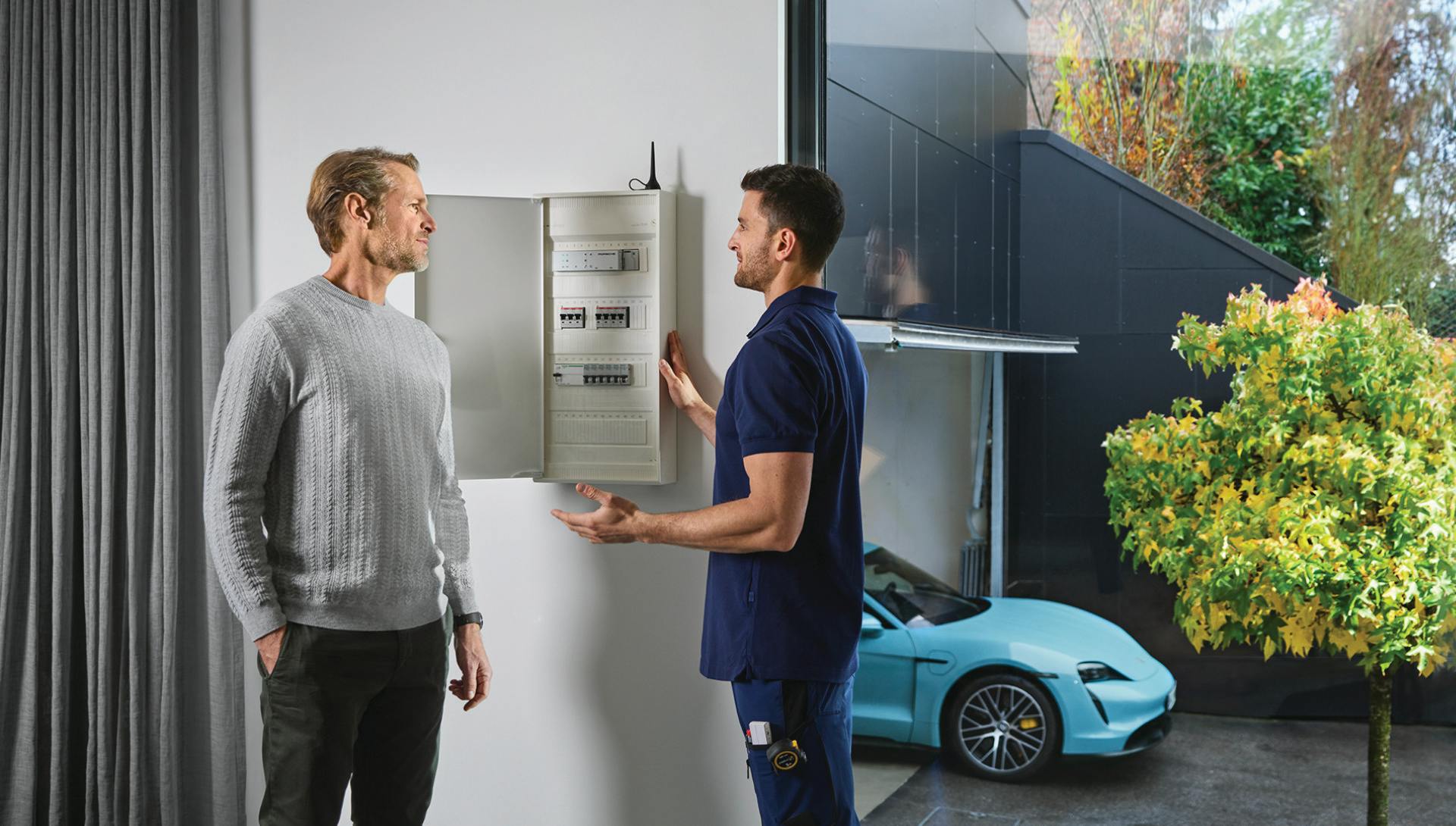 A service technician is standing in a house with a man at an open fuse box. A Porsche Taycan is parked in the background.