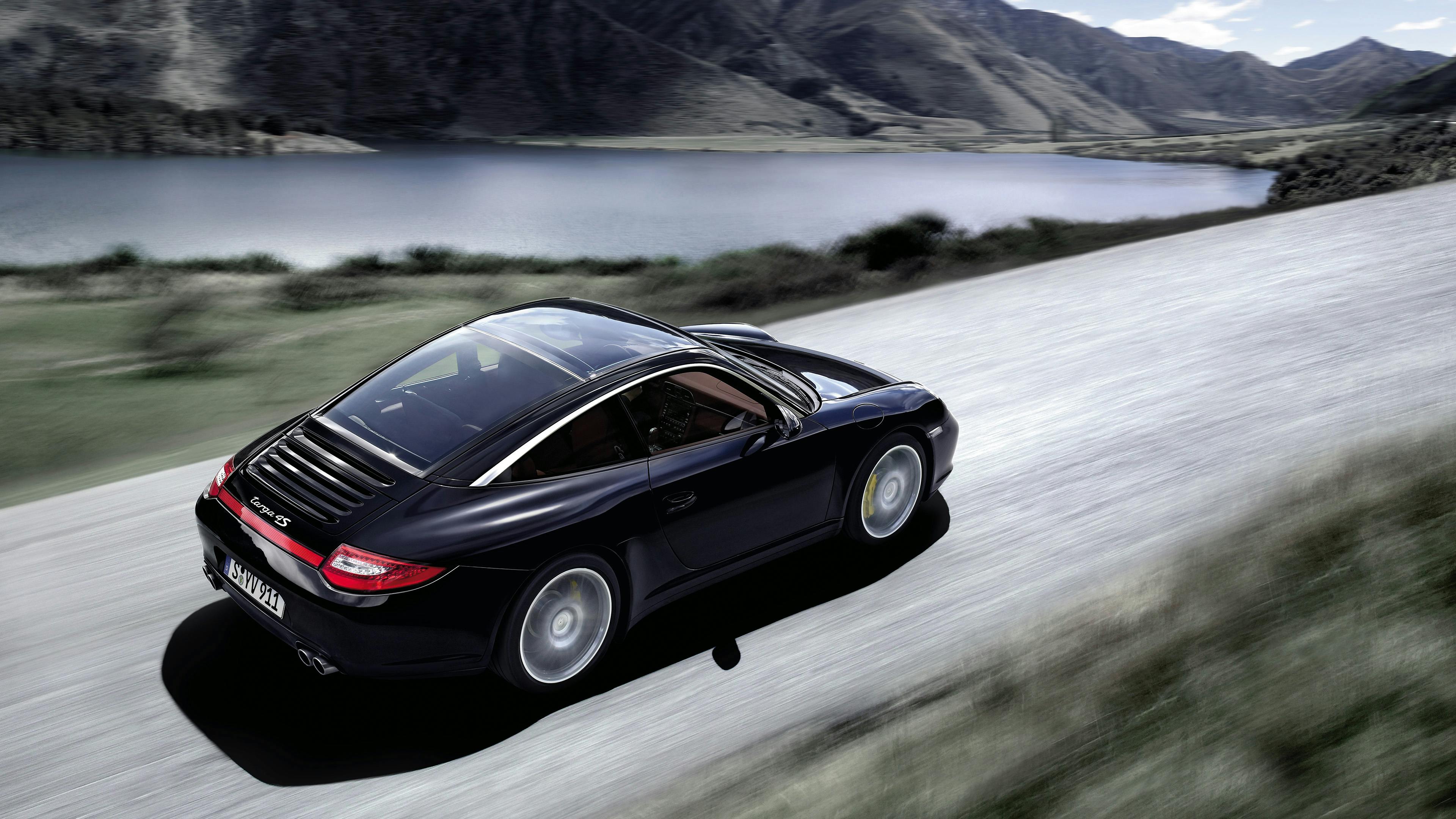 Driving black 997 Targa 4S on a country road by a lake.