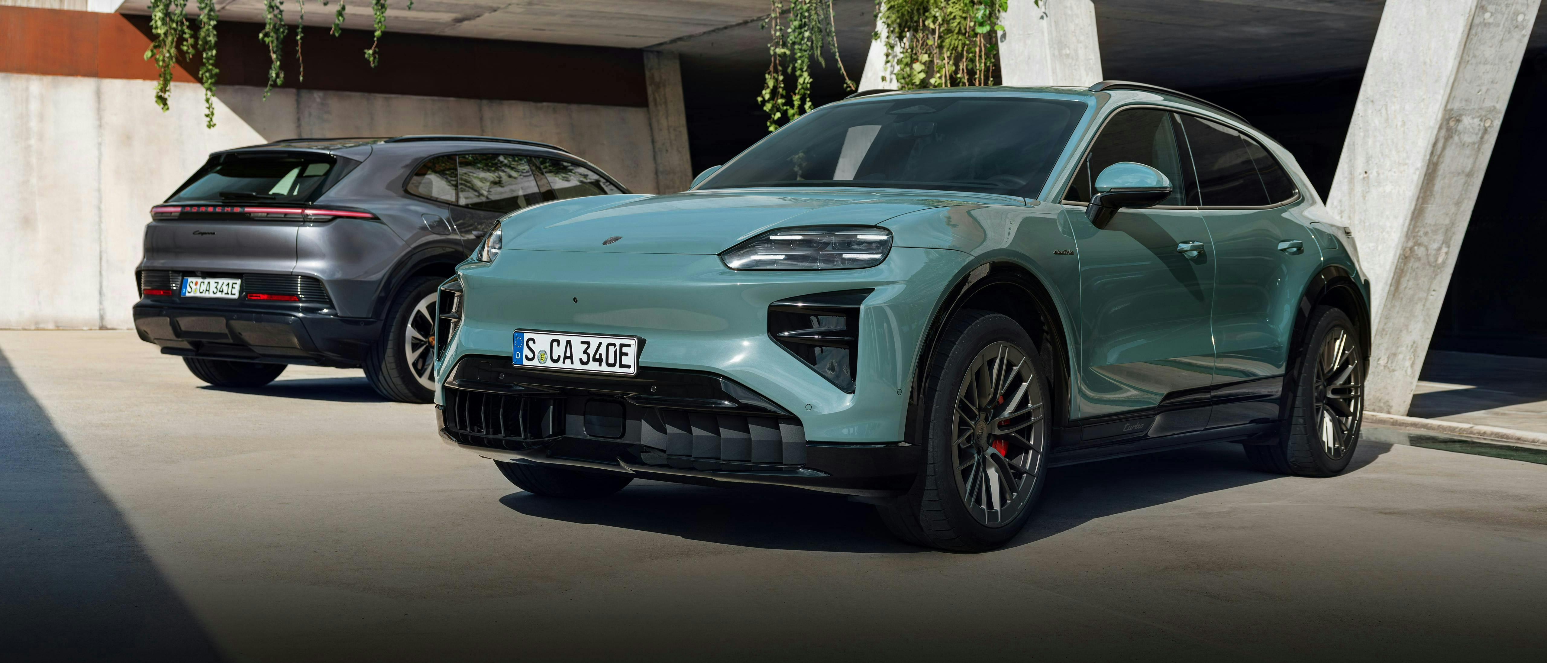 Zwei elektrische Porsche Cayenne stehen auf einem modernen Architekturplatz, vorne in Türkis, dahinter in Grau.
