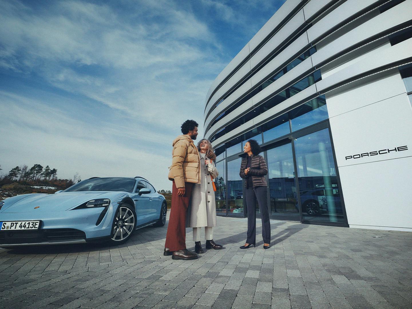 Three people standing next to a blue Porsche Taycan in front of a Porsche dealership.