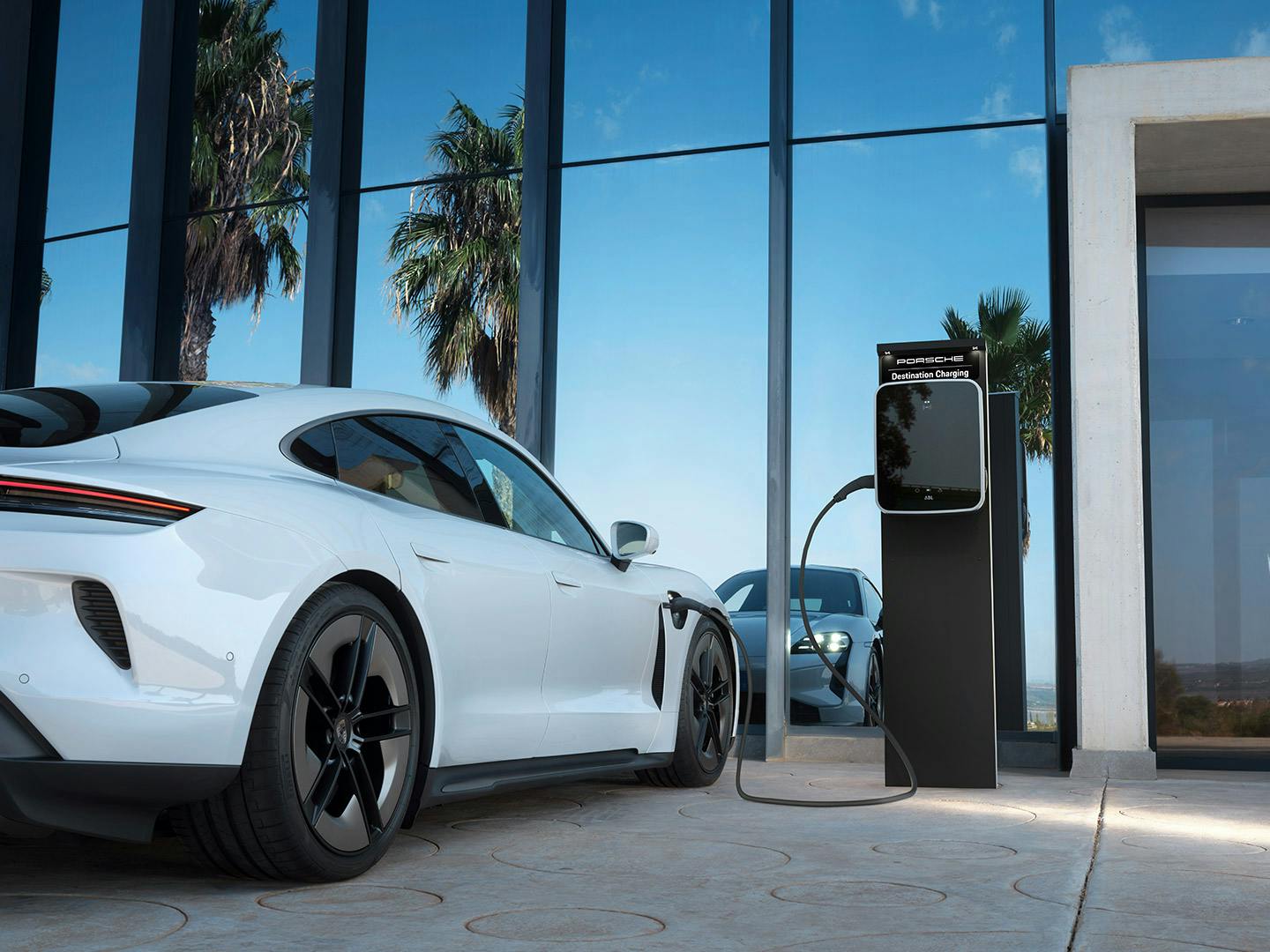Ein weißer Porsche Taycan lädt an einer Porsche Charging-Station vor einem modernen Gebäude mit Glasfassade.