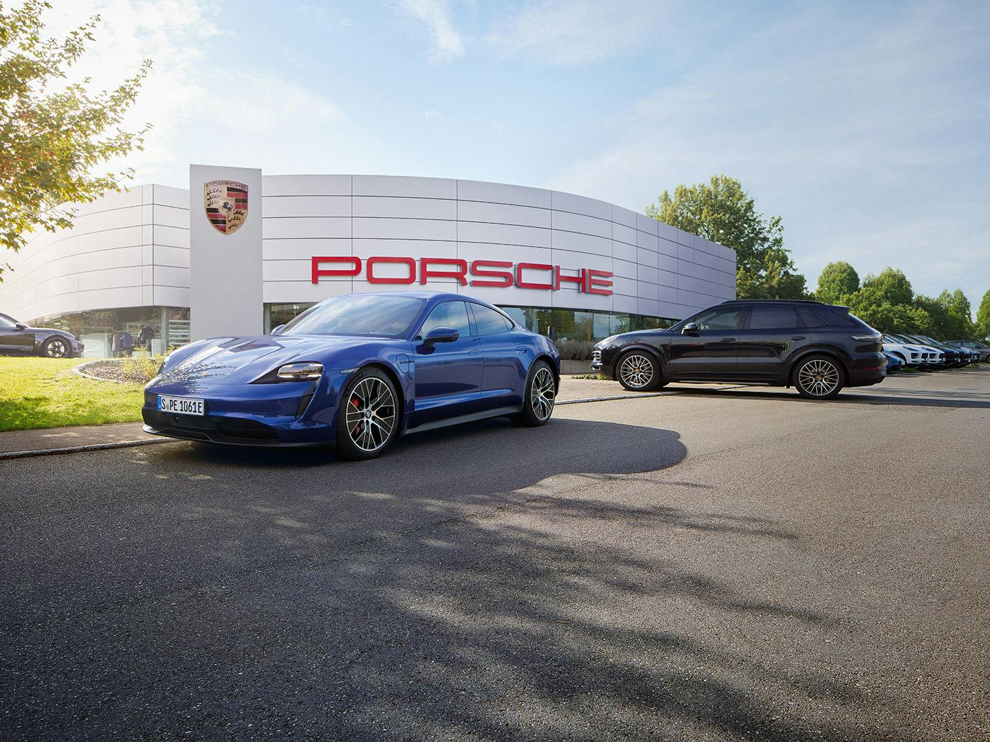 Blauer Porsche Taycan und schwarzer Porsche Cayenne vor einem Porsche-Autohaus  bei sonnigem Wetter.