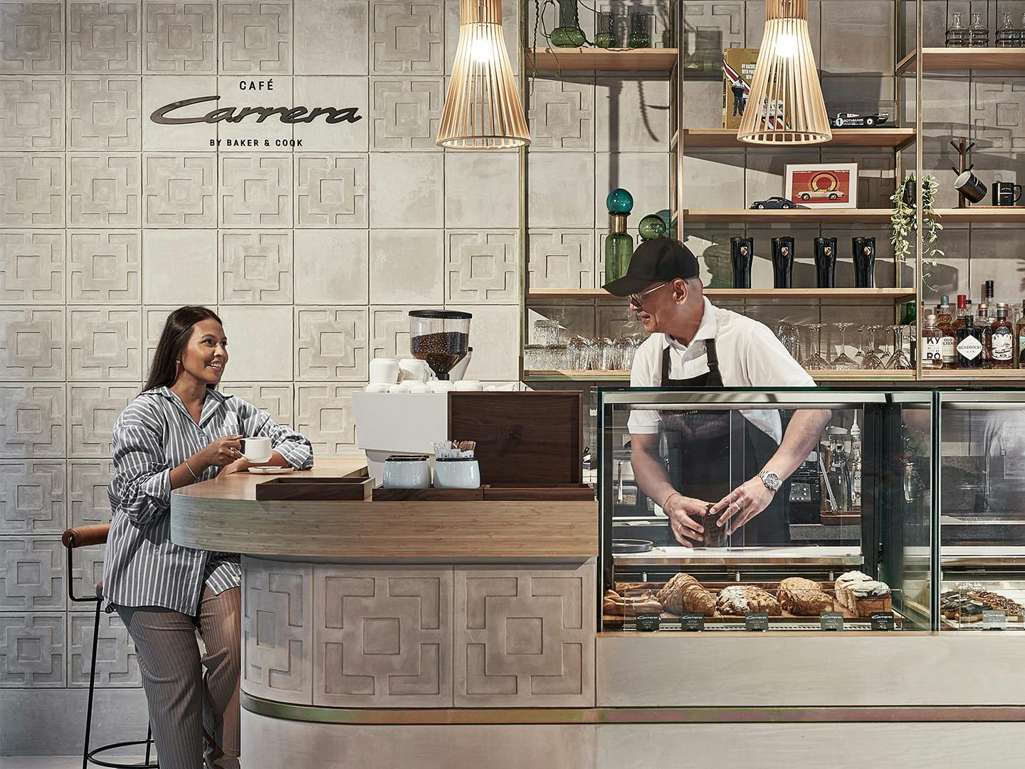 A woman ordering coffee at a stylish café counter labeled “Carrera”, with a barista preparing pastries behind a glass display.