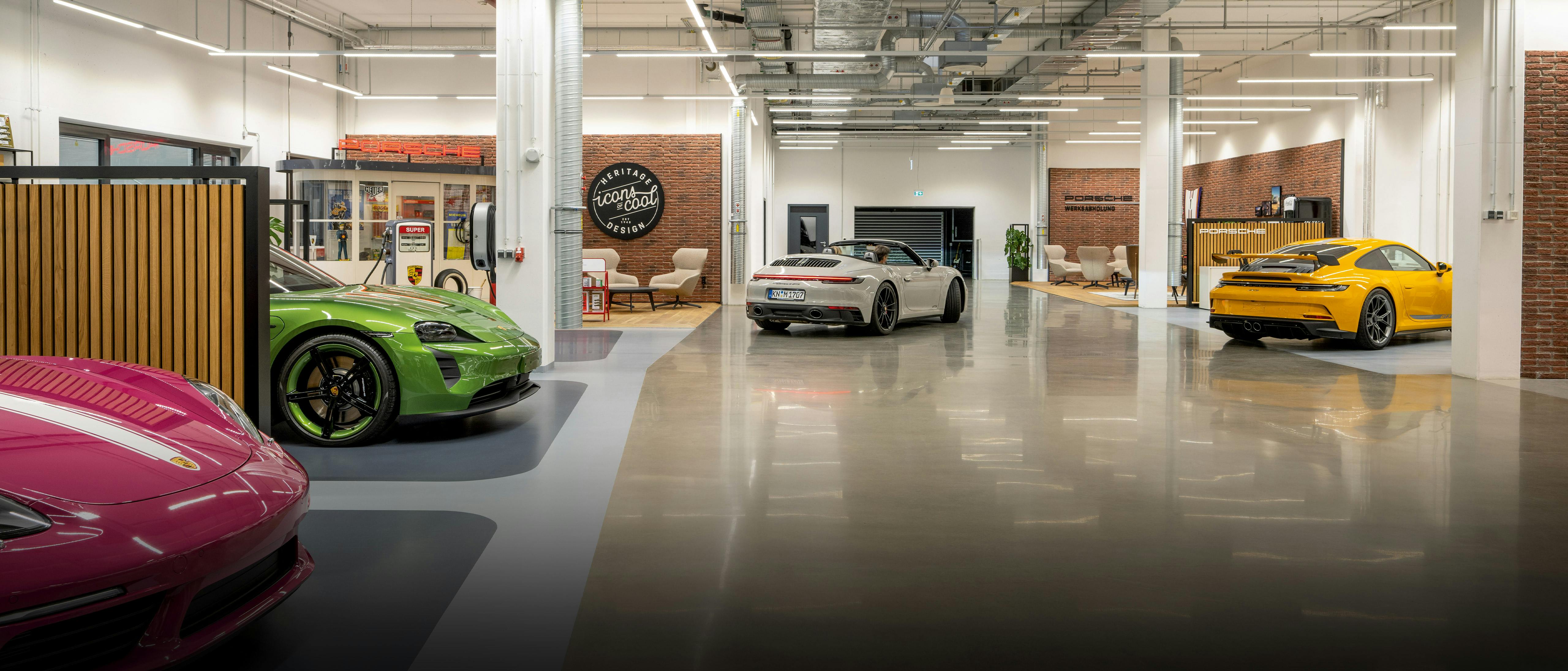 Mehrere Porsche Fahrzeuge stehen verteilt in einer Halle zur Porsche Werksabholung. Am Ende der Halle sind Sitzbereiche mit Sesseln.
