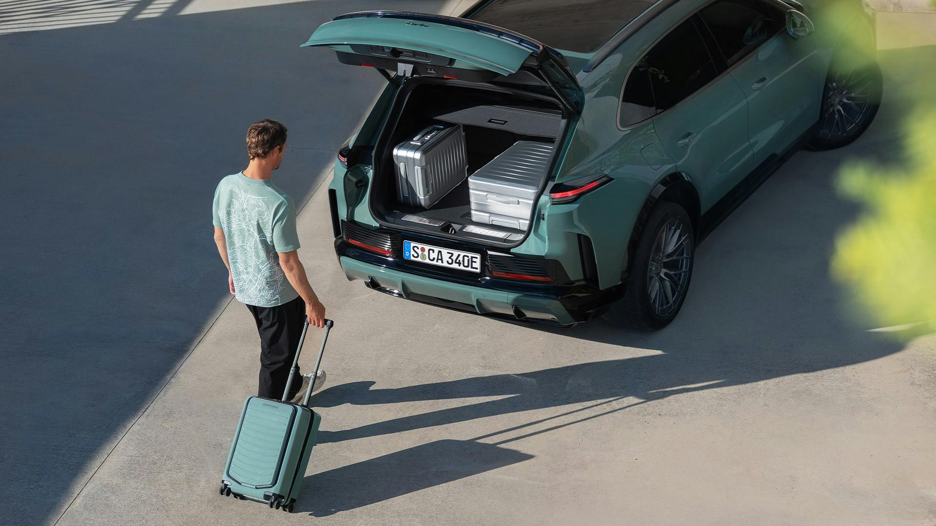 Un hombre con una maleta con ruedas verde Porsche Design camina hacia el maletero abierto de un Cayenne Turbo E-Hybrid, en el que hay guardadas otras dos maletas Porsche plateadas.