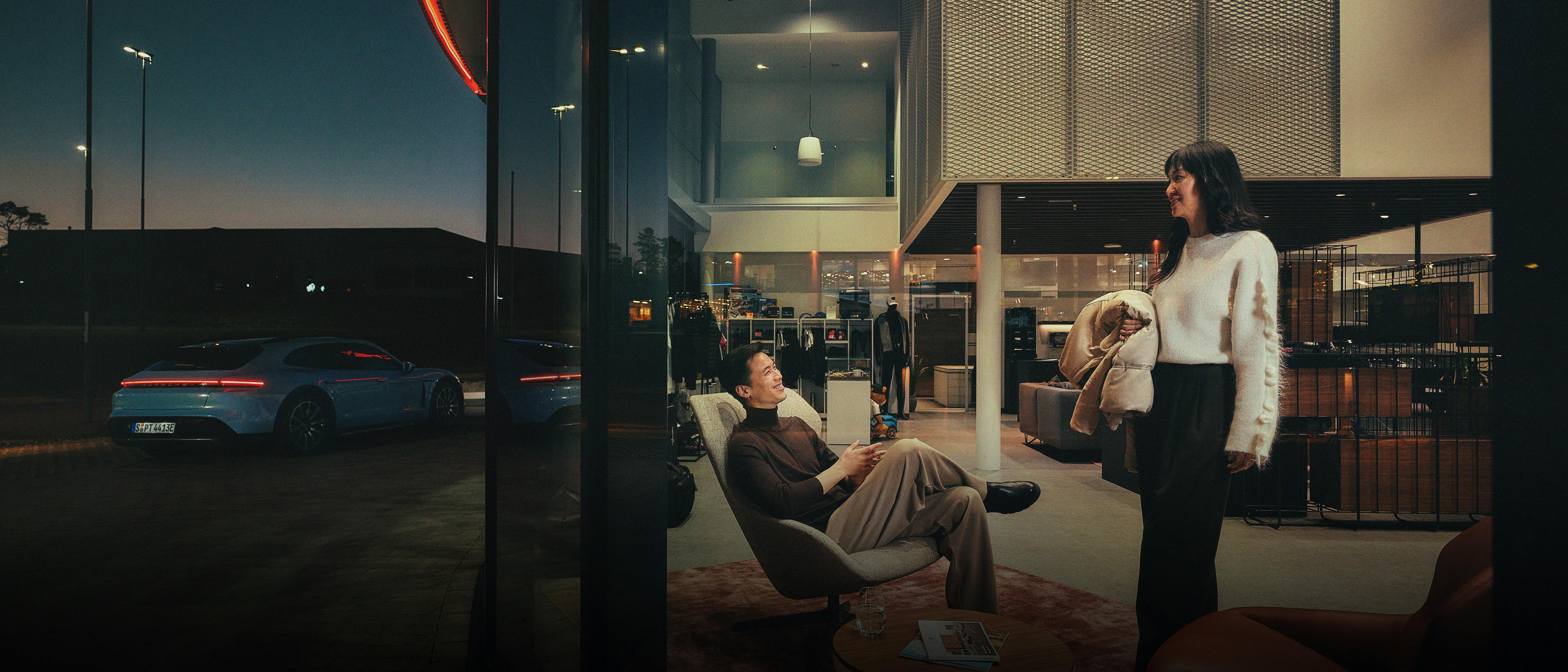 A couple in a Porsche Centre in the evening. The man is sitting on an armchair and the woman is standing next to him holding her coat.