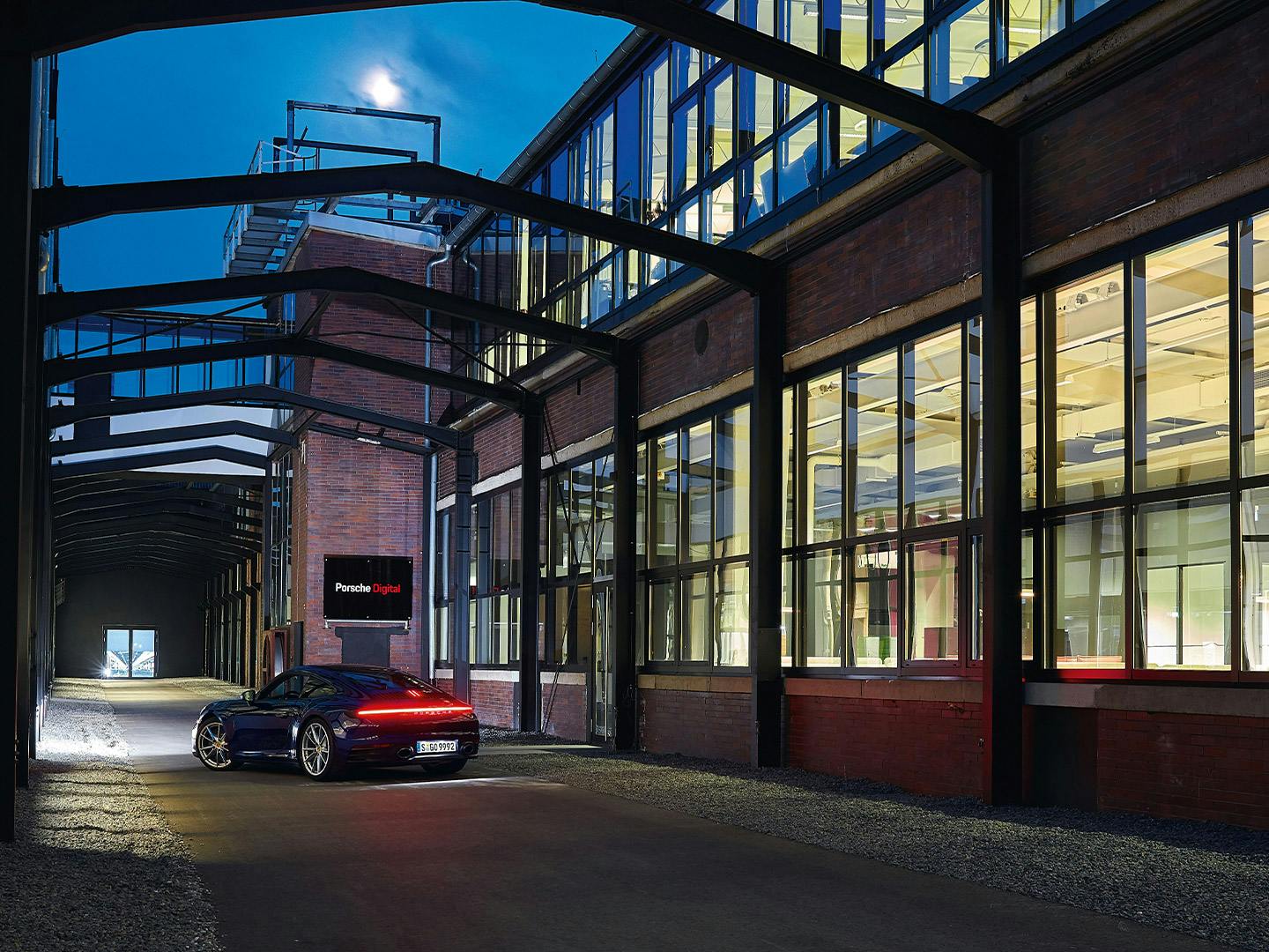 Outdoor hall of a brick industrial building at dusk. A Porsche 911 model is parked in front of the entrance with the Porsche Digital company sign.