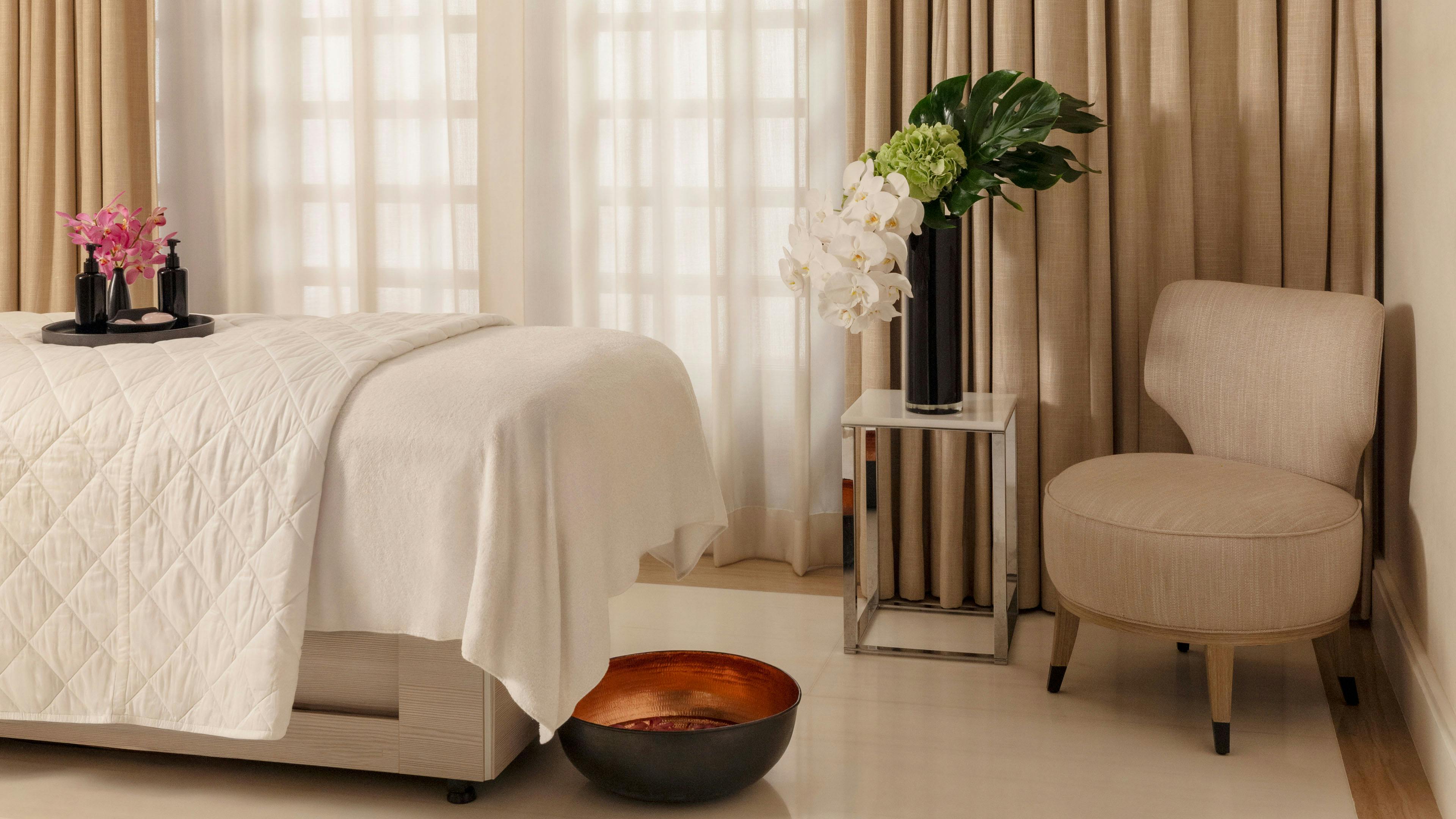 Raffles Hotel spa room with a massage bed, beige curtains, a chair, and flowers on a side table.