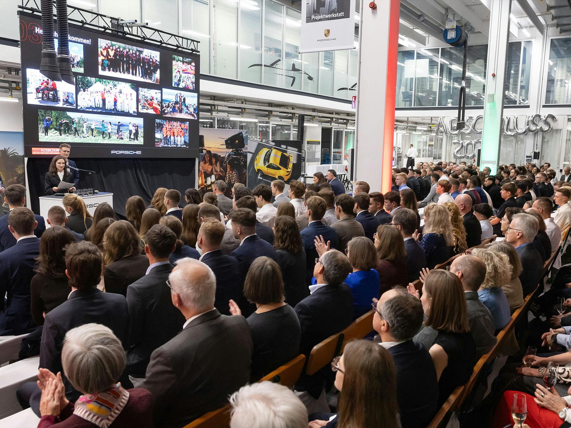 Großes Porsche Event in einem modernen Industrieumfeld: Publikum sitzt in Reihen und verfolgt eine Präsentation vor einer großen LED‑Leinwand mit Team‑ und Projektbildern.