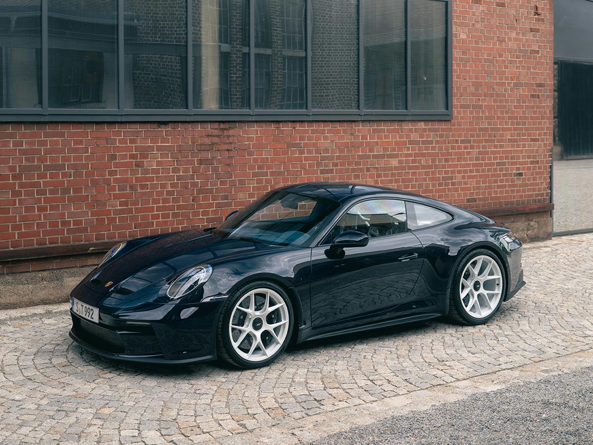 Dark blue Porsche 911 on cobblestone pavement in front of brick facade with large industrial-style windows.