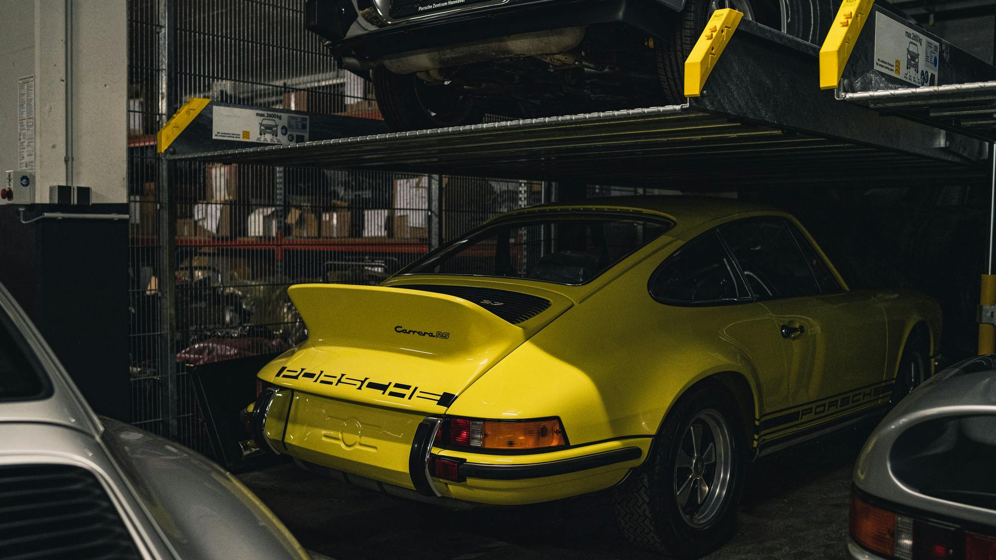 A bright yellow vintage Porsche Carrera RS parked inside a multi-level vehicle storage facility or garage. The car is positioned under a mechanical car lift holding another vehicle above it. The rear of the yellow car is clearly visible, showcasing its iconic "ducktail" spoiler and black Porsche lettering. Parts of other silver cars frame the foreground.