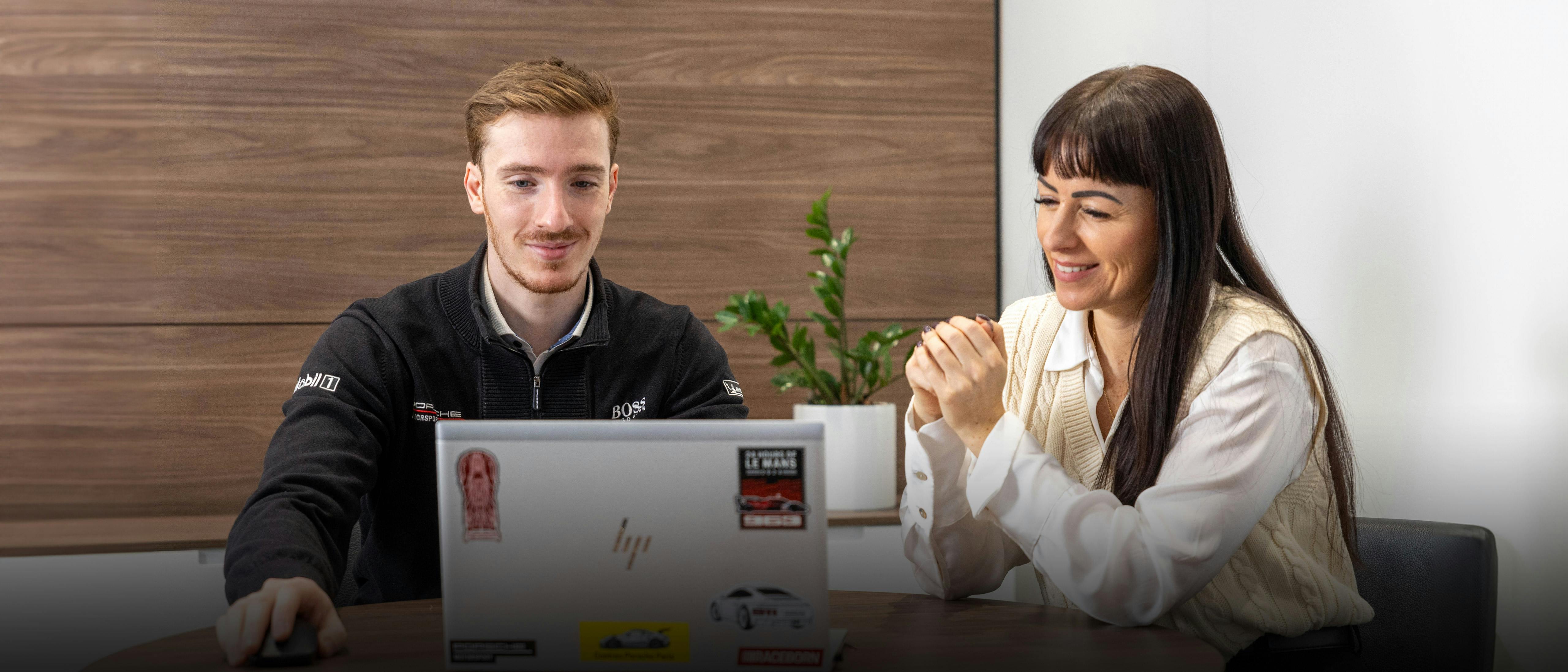 Un conseiller Porsche montre à une femme quelque chose sur un ordinateur portable gris avec des autocollants Porsche. Tous deux sont assis à une table en bois et ont l'air sympathique.