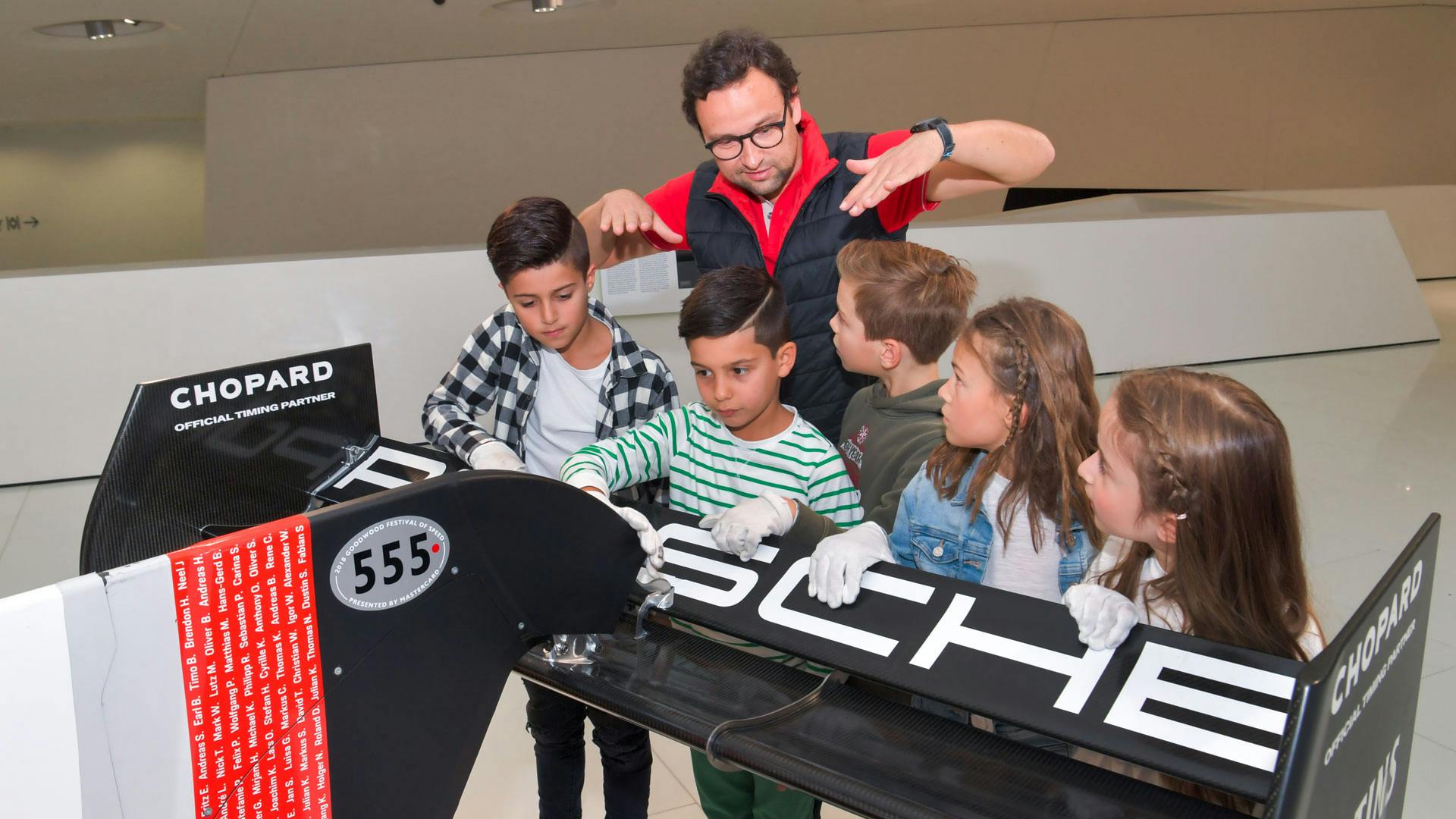 Eine Gruppe von Kindern und ein Porsche Museum Mitarbeiter, die am Heck eines Autos stehen.