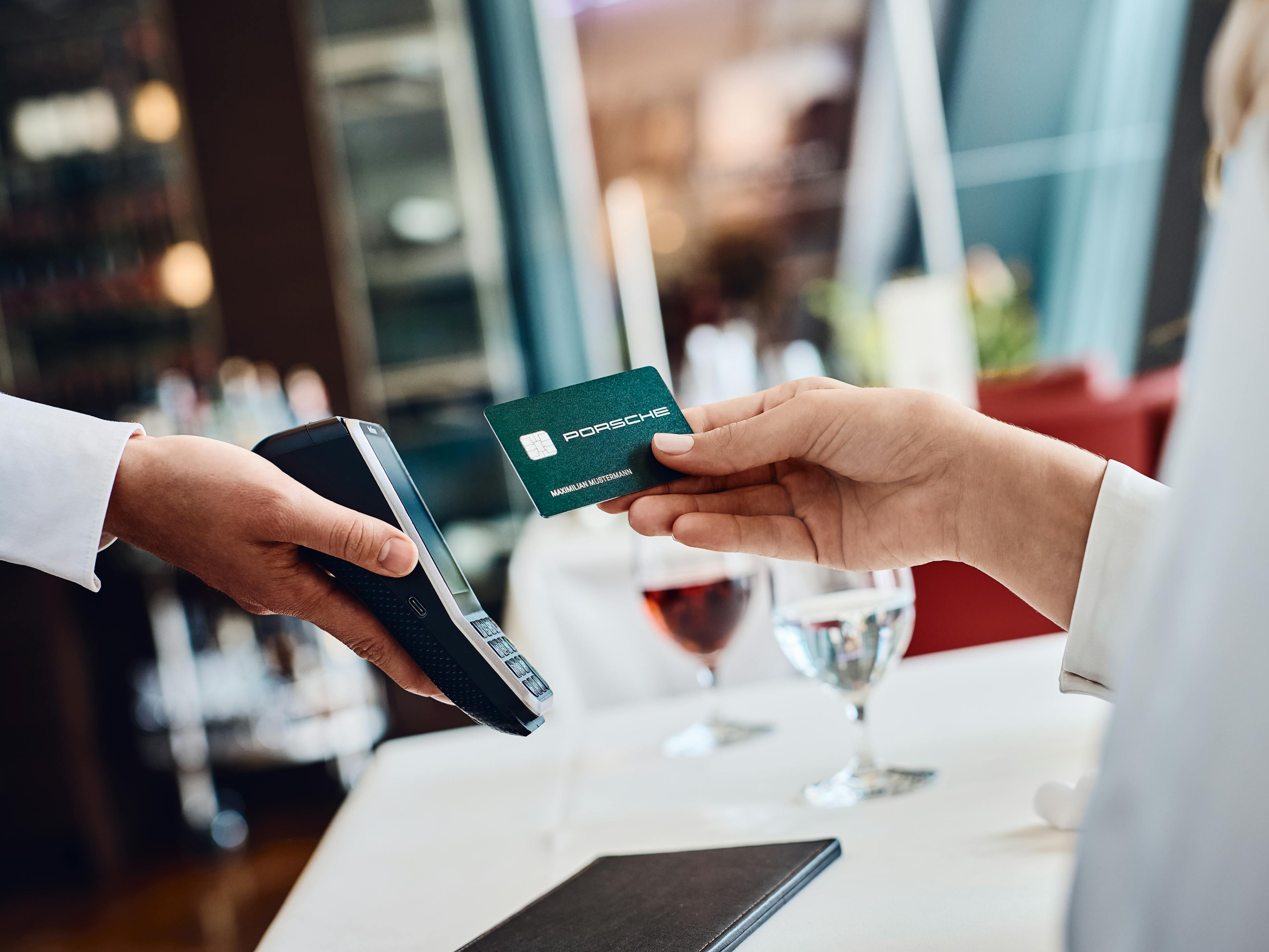 Eine Person hält eine Porsche-Kreditkarte über ein Kartenlesegerät, um eine Zahlung in einem Restaurant durchzuführen. Auf dem Tisch stehen zwei Gläser mit Getränken und eine schwarze Rechnungsmappe.