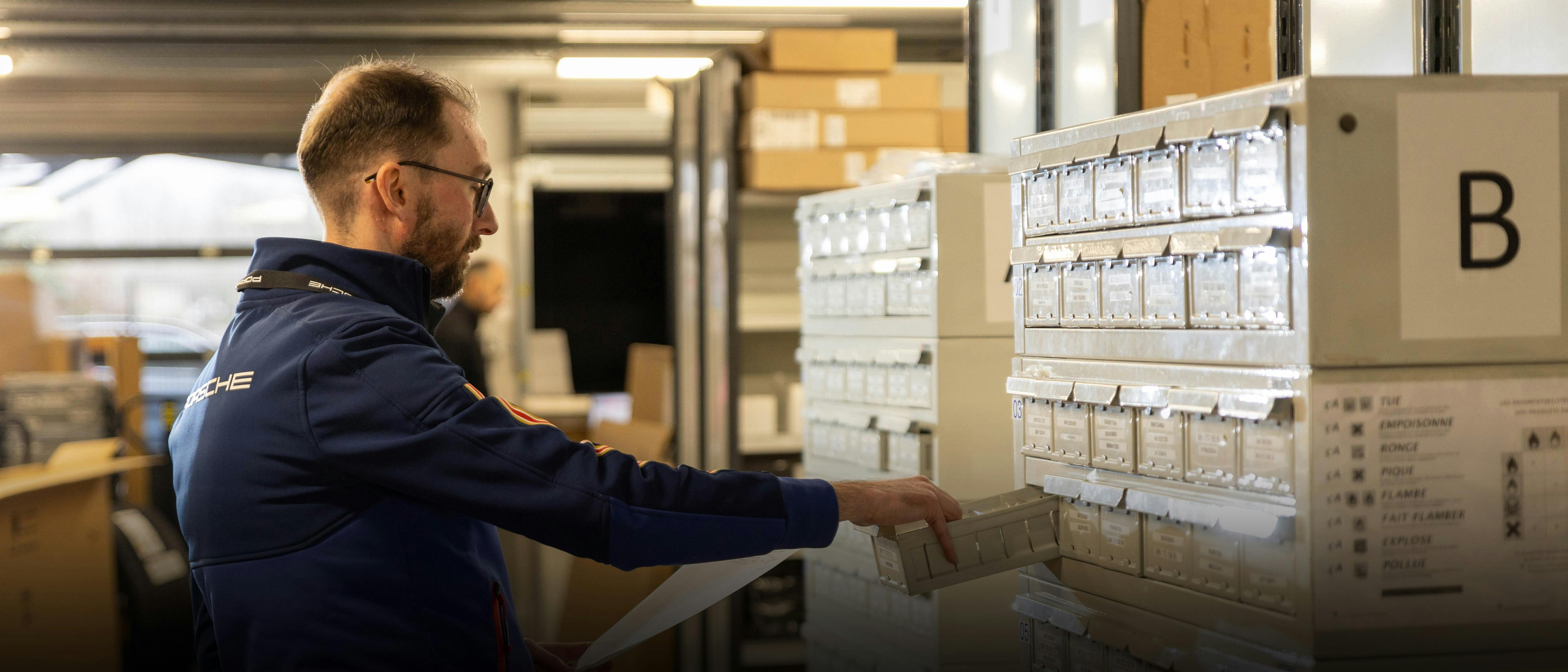 Un employé de Porsche Logistics en tenue de travail se tient devant une armoire à tiroirs, tire un tiroir et tient des documents dans l'autre main.