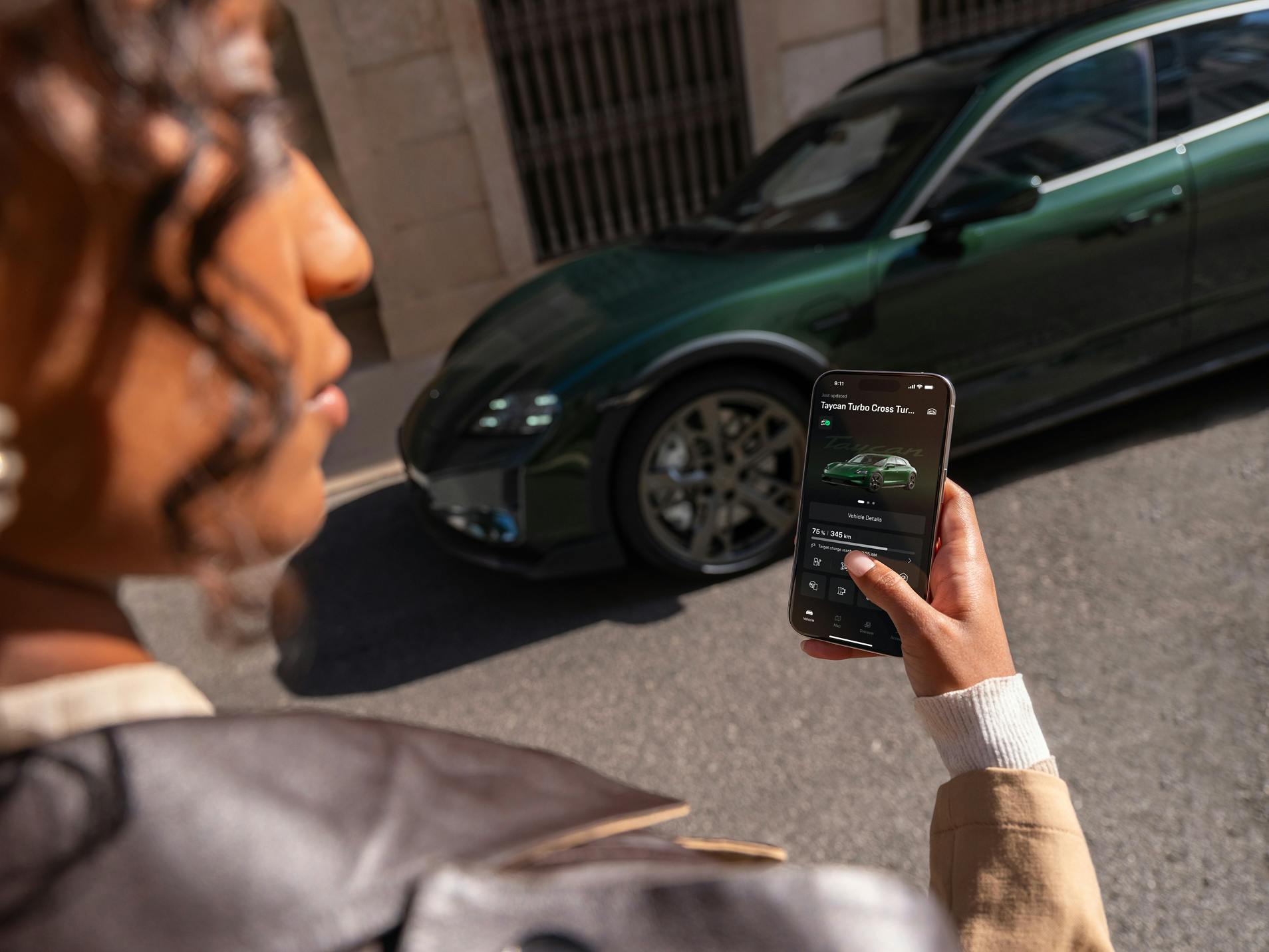 A woman looking at her smartphone with the My Porsche App open. A Porsche Taycan Turbo Cross Turismo is in the background.