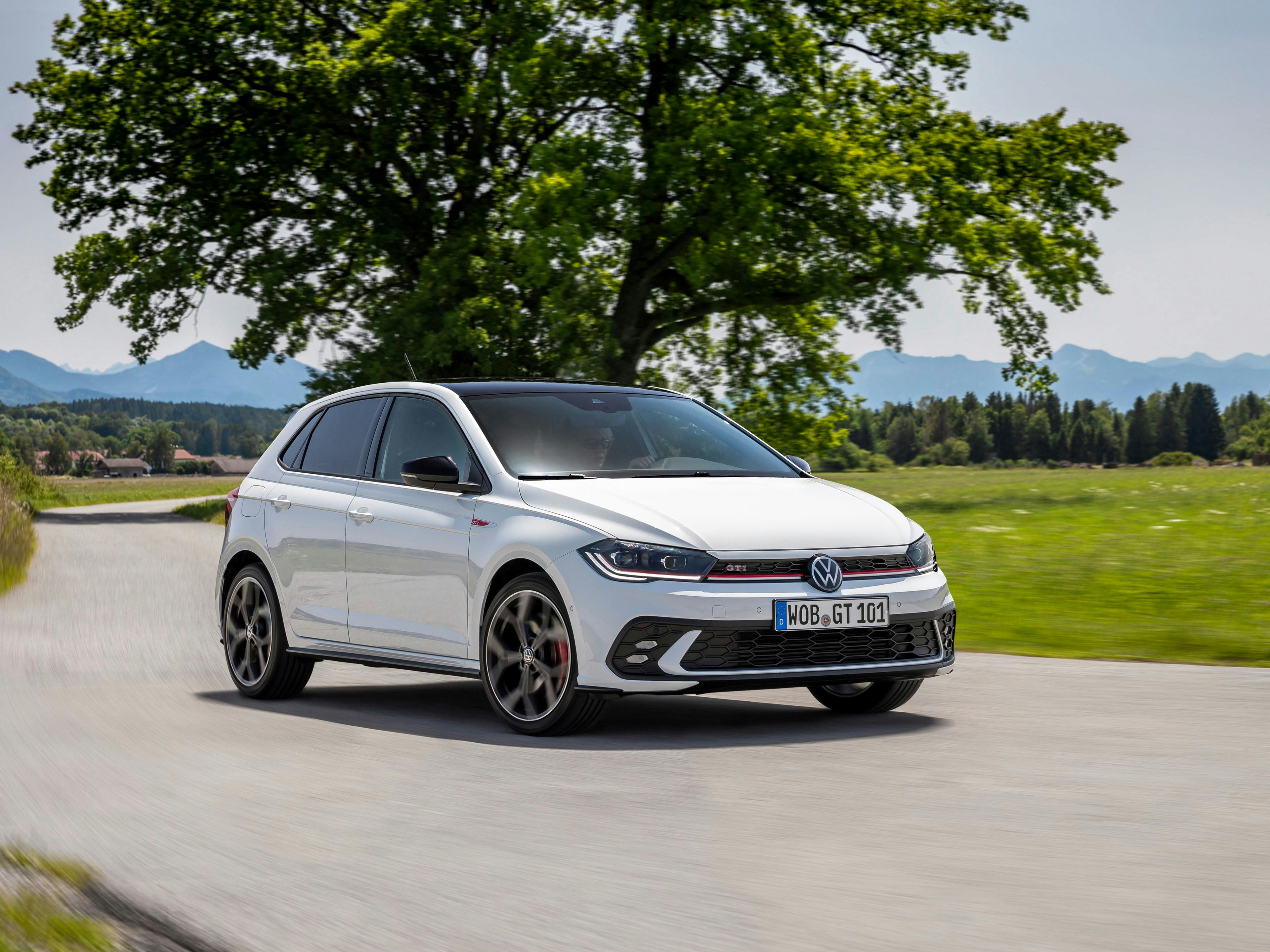 Ein weißer VW Polo GTI fährt auf einer Landstraße, Bäume und Wiesen im Hintergrund.