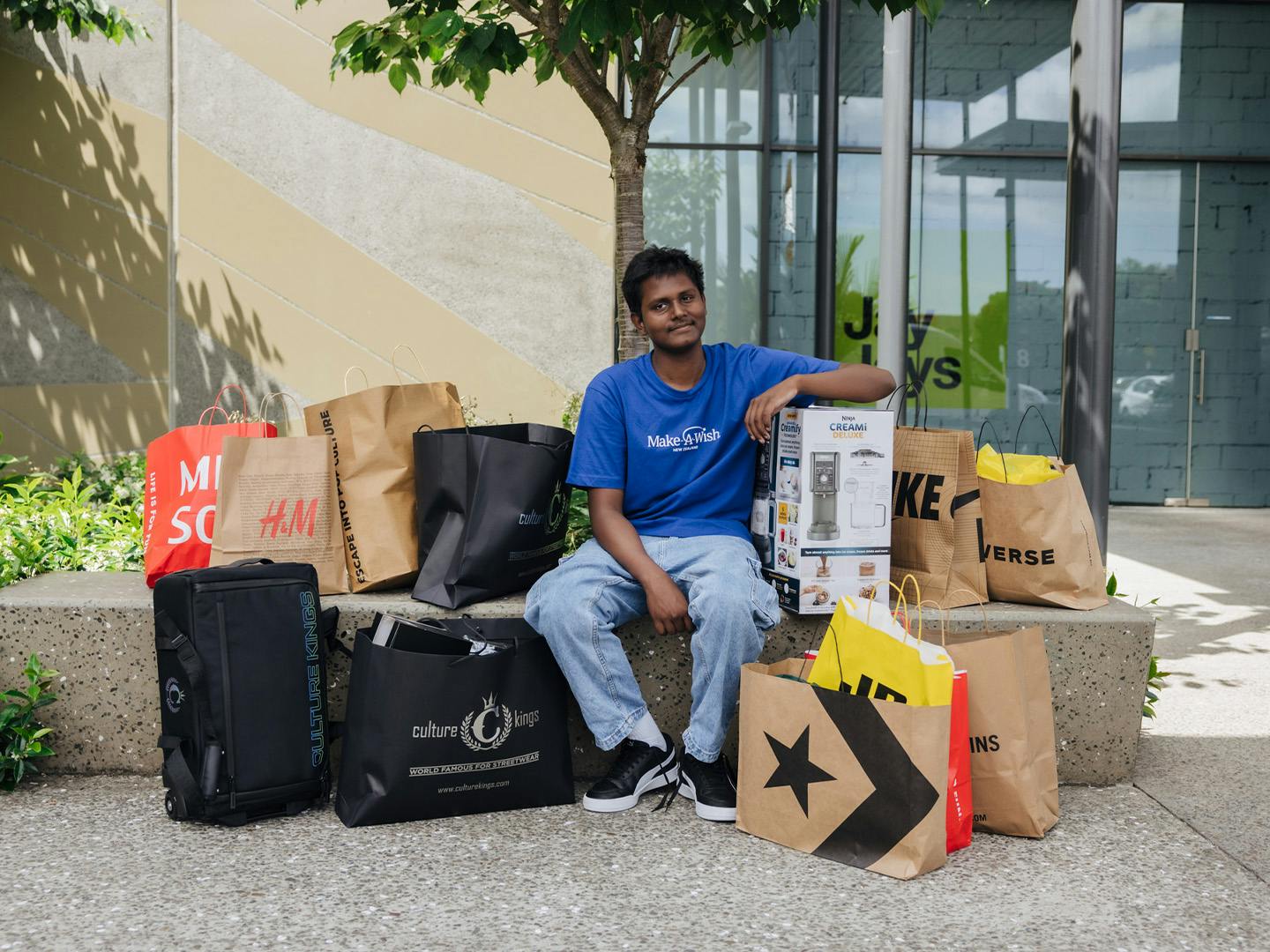 A teenage boy sitting outdoor, smiling, surrounded by numerous shopping bags and boxes from various brands. He looks content and is resting his arm on a new item from his shopping spree.