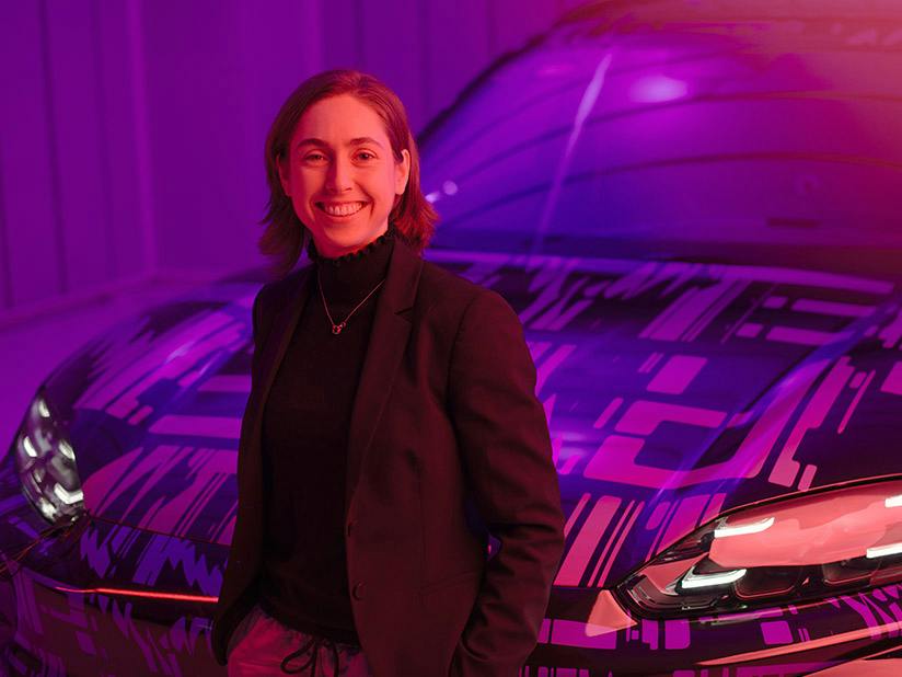 Porsche Mitarbeiterin steht vor einem sportlichen Fahrzeug mit abstraktem Design; violette Beleuchtung und moderner Studiohintergrund.