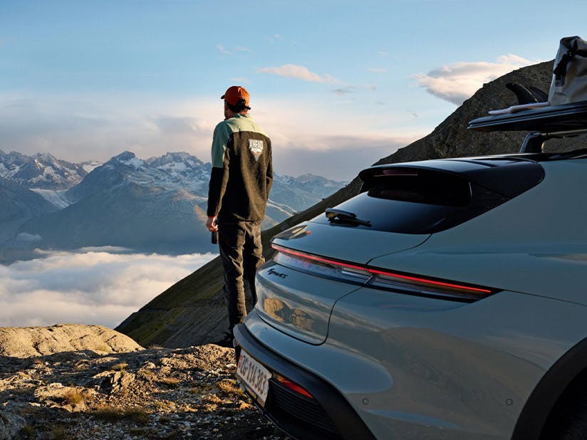 Person in outdoor gear standing beside silver Porsche Taycan Cross Turismo, overlooking dramatic alpine mountain landscape with clouds in valley below