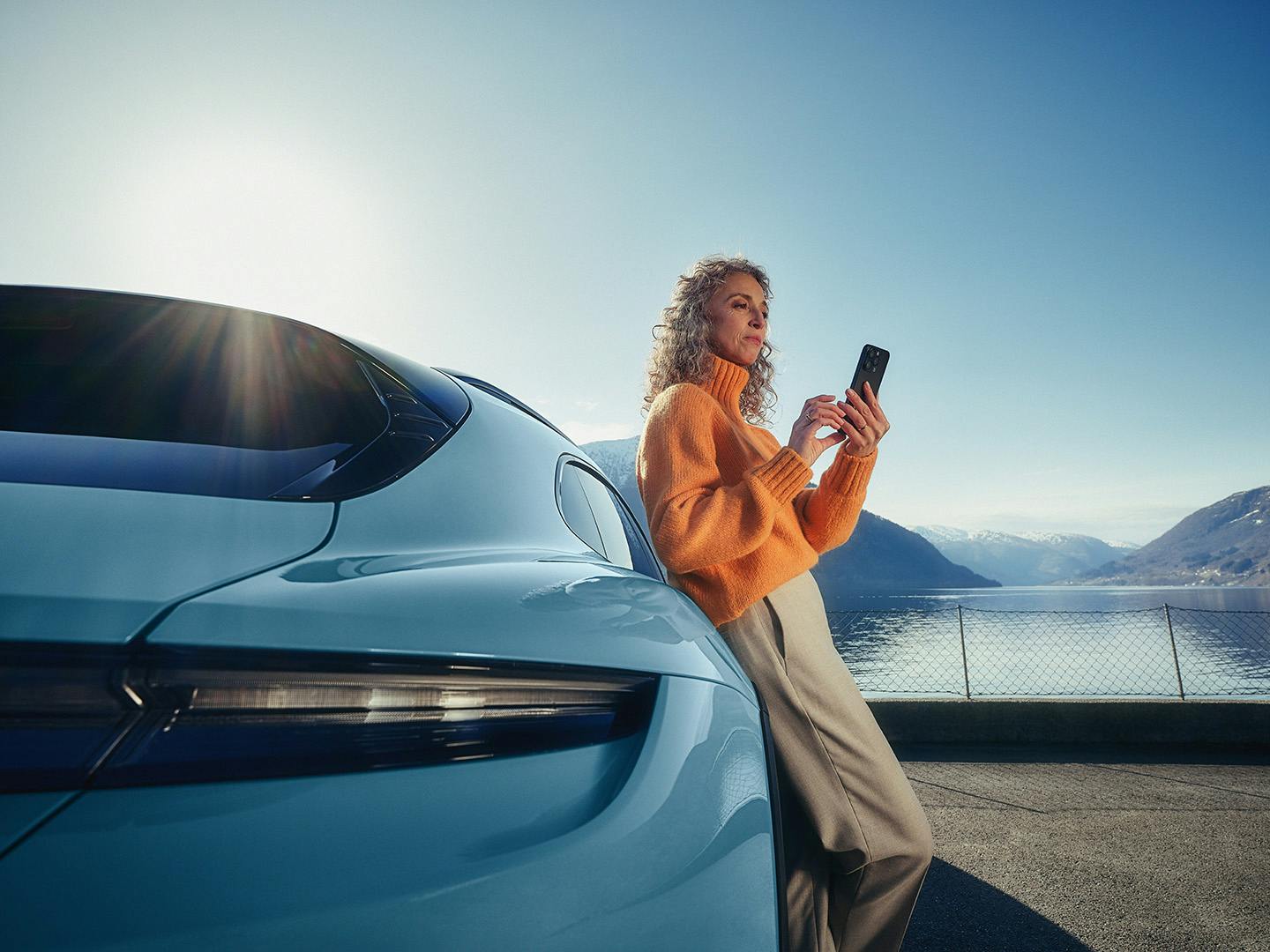 A female customer leaning on a car while using her phone. Mountains and sea can be seen in the background.