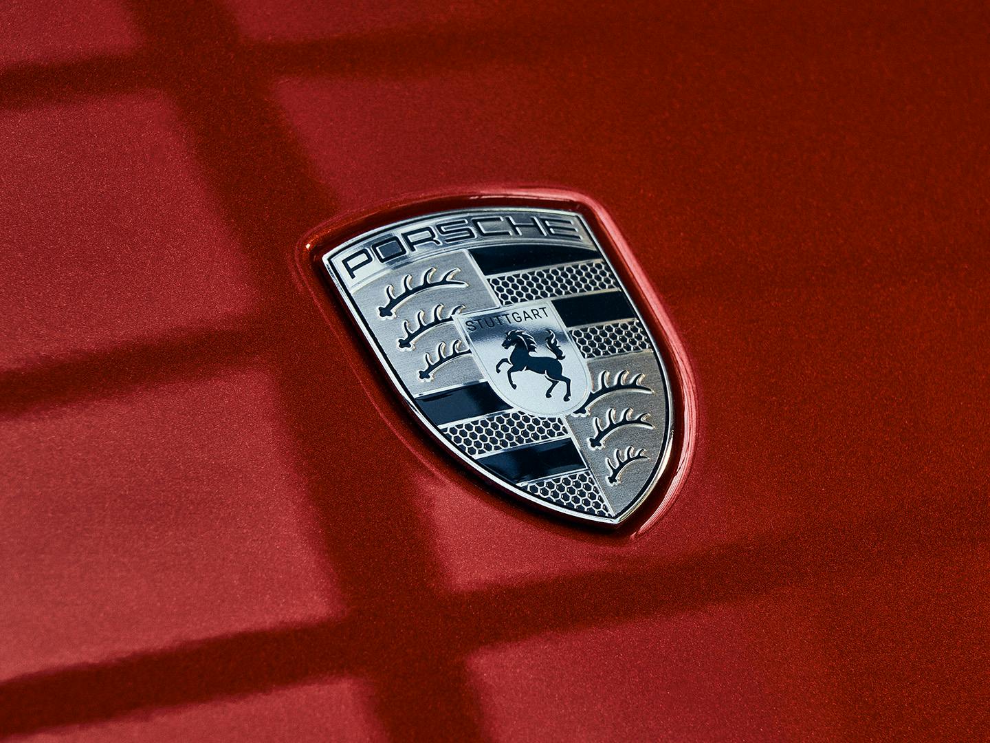 A close-up of the Porsche logo on a red car. The emblem features the word "Porsche" at the top, a black prancing horse in the center.