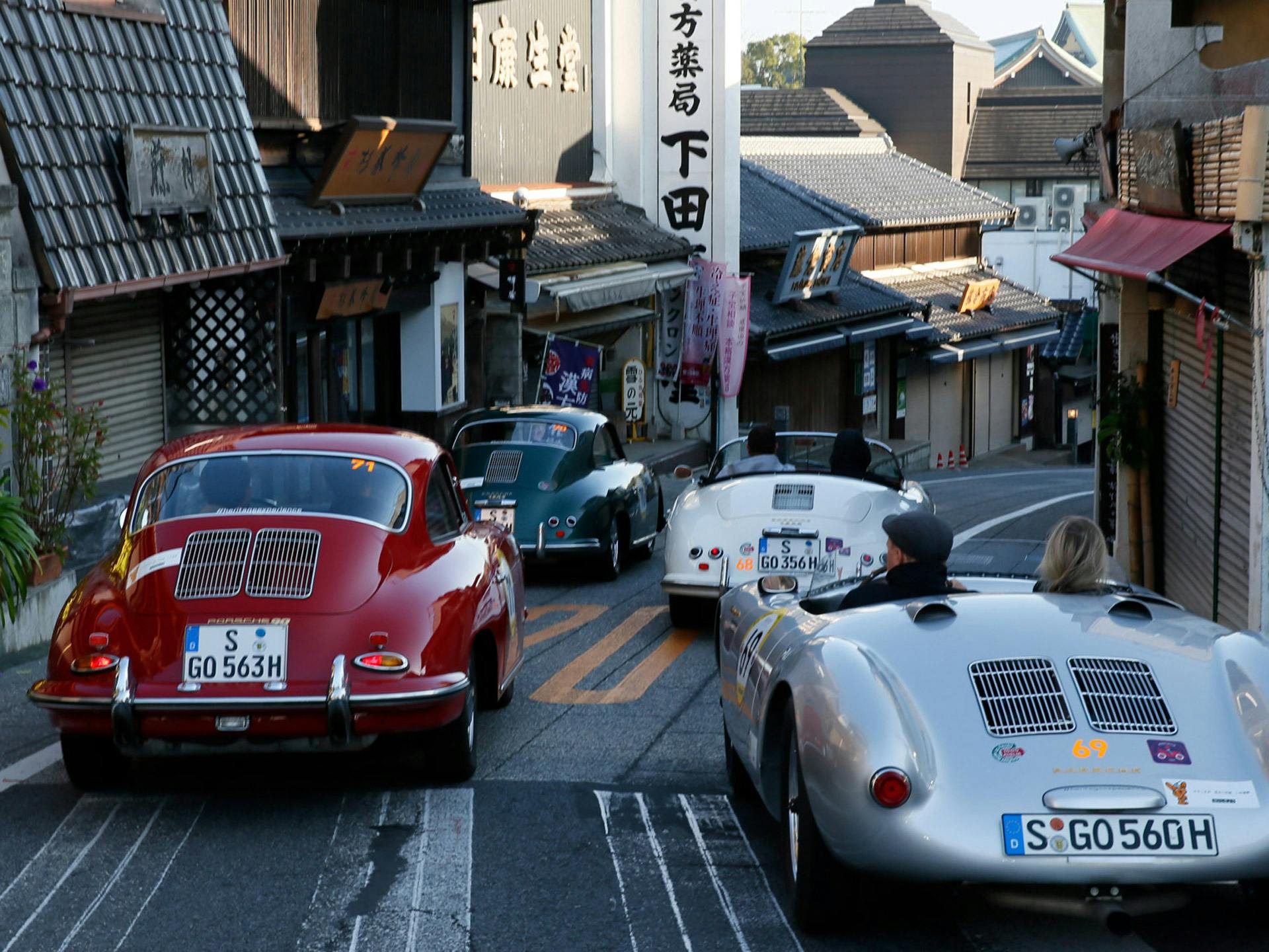 Mehrere verschiedenfarbige klassische Porsche Fahrzeuge die um eine Kurve in einer kleinen Stadt fahren