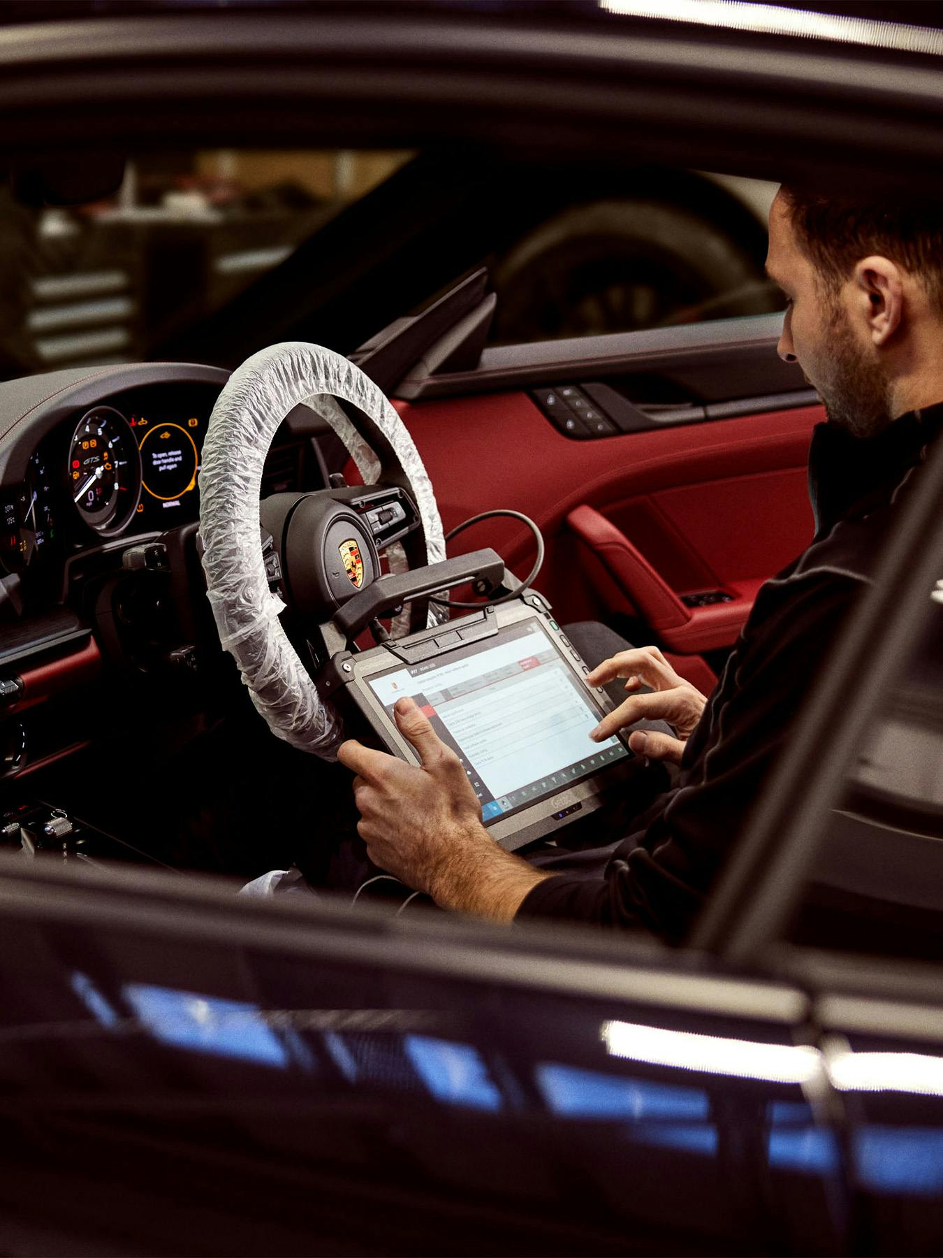 A technician sits in the driver’s seat of a Porsche, which features a red and black interior. The steering wheel is covered in protective plastic. He is holding a large industrial tablet connected to the car's system. The view is from outside the passenger window.