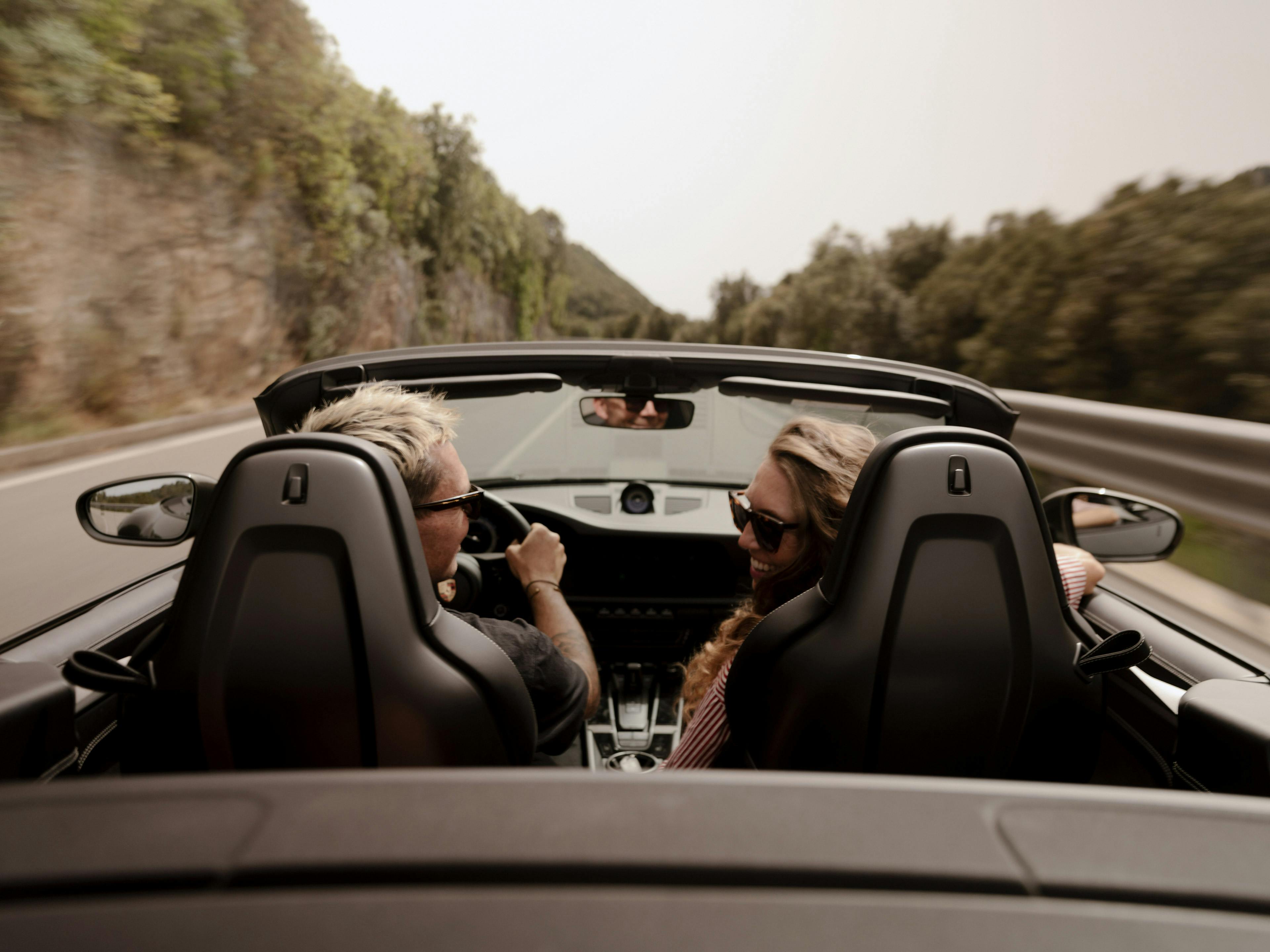 Zwei Personen fahren in einem schwarzen Cabrio mit offenem Verdeck eine kurvige Straße entlang, umgeben von Felsen und Bäumen. Beide tragen Sonnenbrillen, lachen sich an und genießen die Fahrt bei sonnigem Wetter. Die Perspektive zeigt den Innenraum von hinten, mit Blick auf das Cockpit und die Straße.