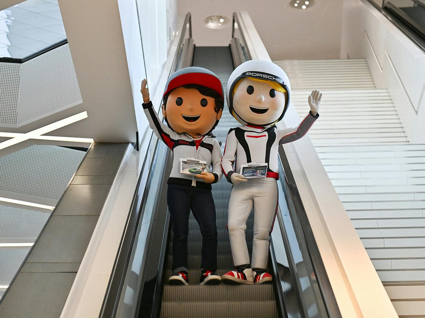 Maskottchen Tom Targa und Tina Turbo grüßen auf der Rolltreppe in die Ausstellung mit dem Entdeckerbuch in der Hand.