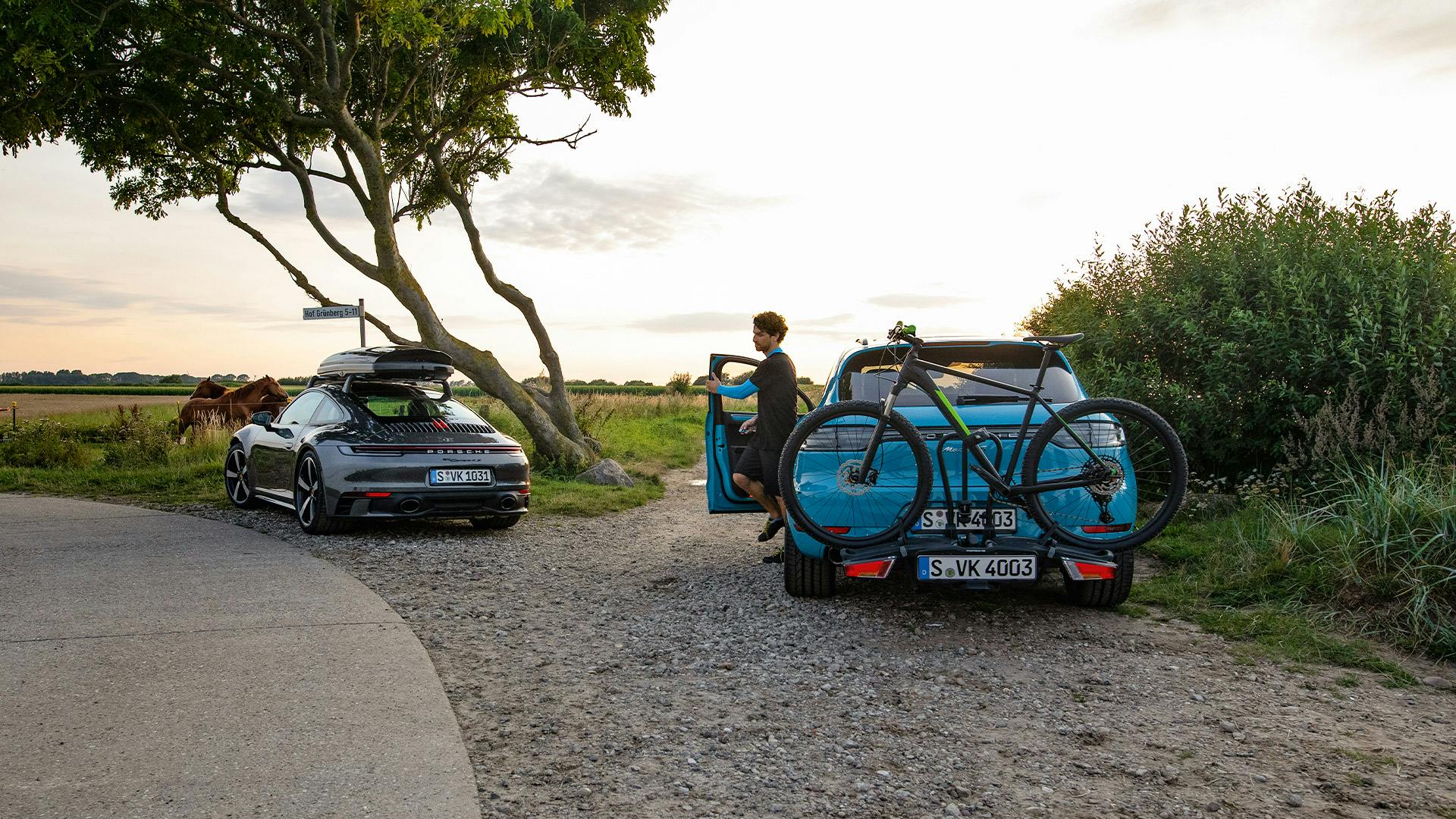 Deux Porsche sont garées dans un environnement rural. L'une d'elles a un coffre de toit et l'autre un porte-vélo avec un vélo dessus.