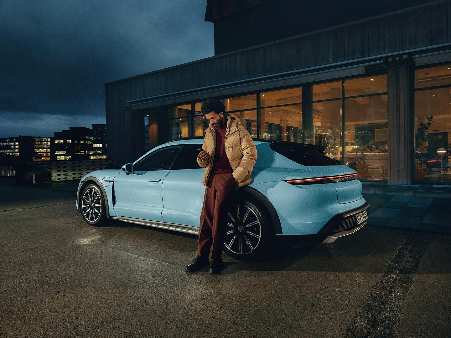 Homme appuyé contre une Porsche Taycan bleu clair, téléphone portable à la main, devant un bâtiment moderne au crépuscule.