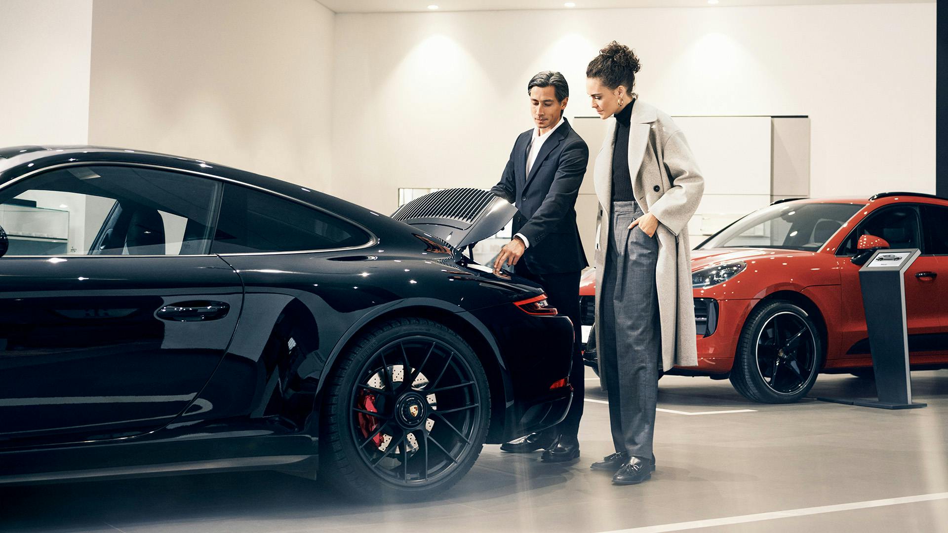 Porsche sales consultant showing vehicle features to a female customer in a modern showroom with black and red Porsche models.