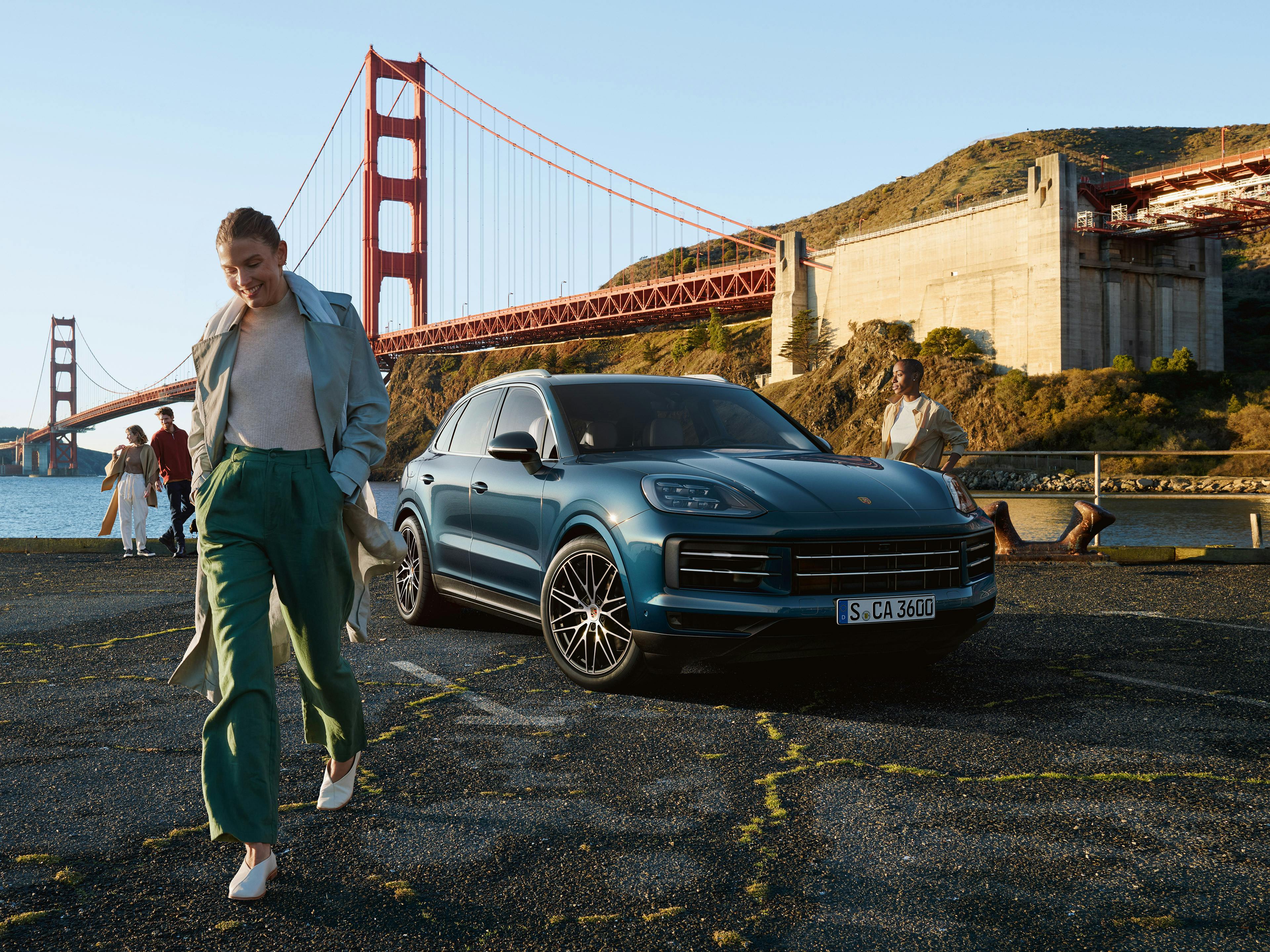 Eine Frau lächelt beim Gehen nahe einem blauen Porsche Cayenne, während die Golden Gate Bridge im Hintergrund zu sehen ist.
