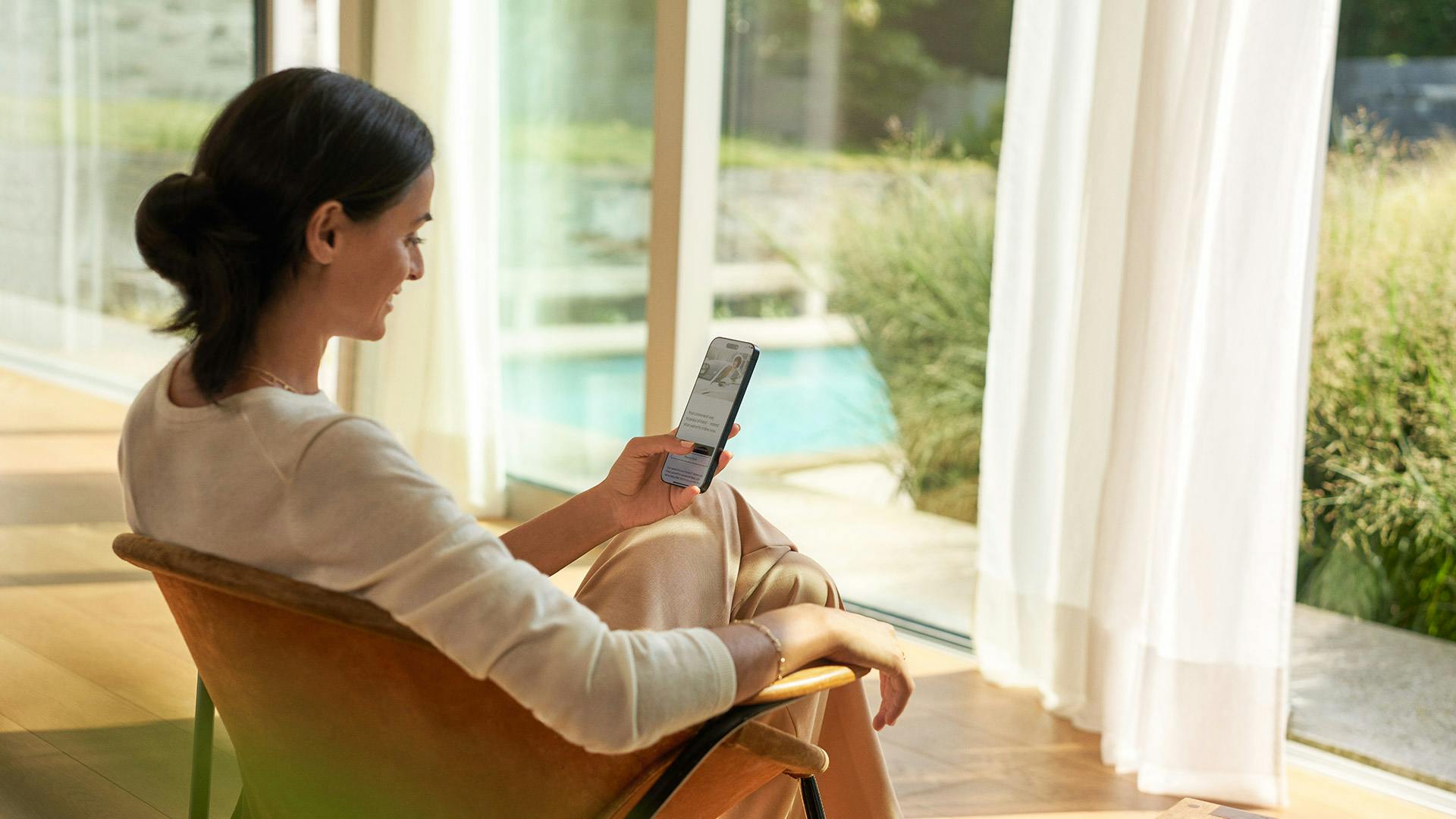 Person sitzt auf einem Stuhl in einem hellen, modernen Raum und nutzt ein Smartphone. Im Hintergrund sind große Fenster mit Blick auf einen Garten oder Außenbereich.