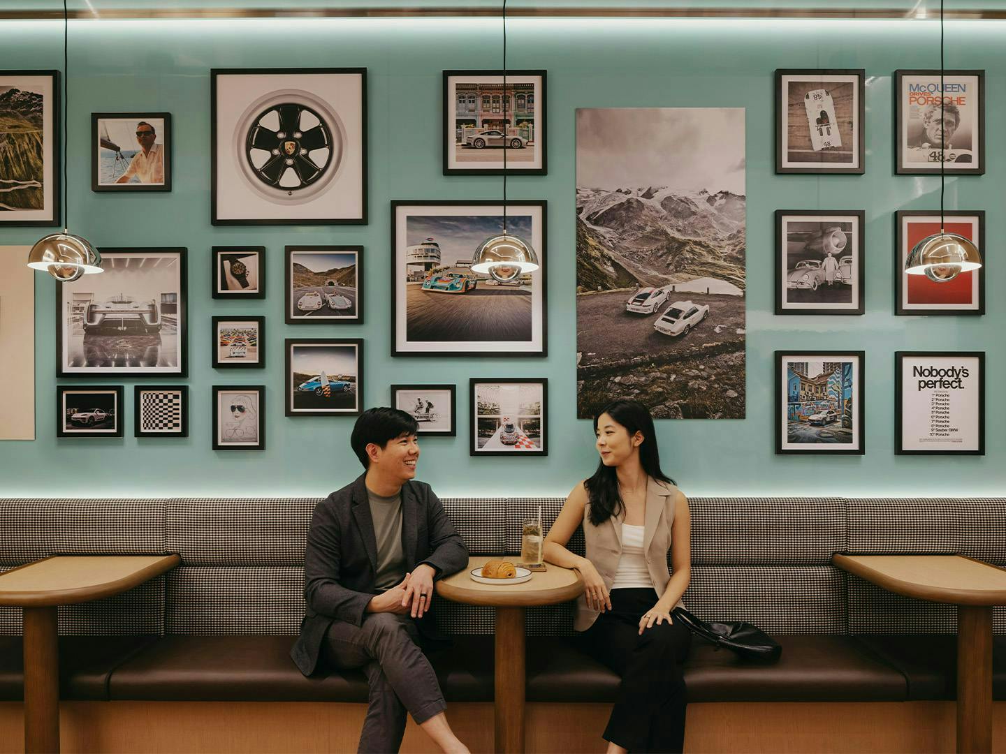 Two guests sit at Café Carrera tables, chatting in front of a gallery wall with Porsche-themed photos and artwork.