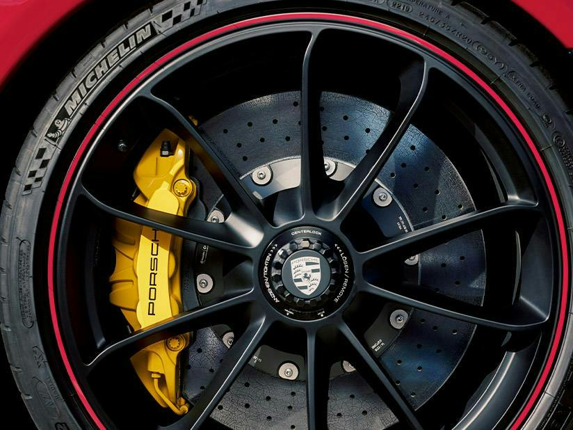 A close-up shot of a black Porsche wheel featuring a centre-lock nut with the Porsche crest. Behind the thin spokes are cross-drilled ceramic brake discs and a large, bright yellow brake calliper branded with the word "PORSCHE." The tire is a Michelin with a thin red accent line around the rim.