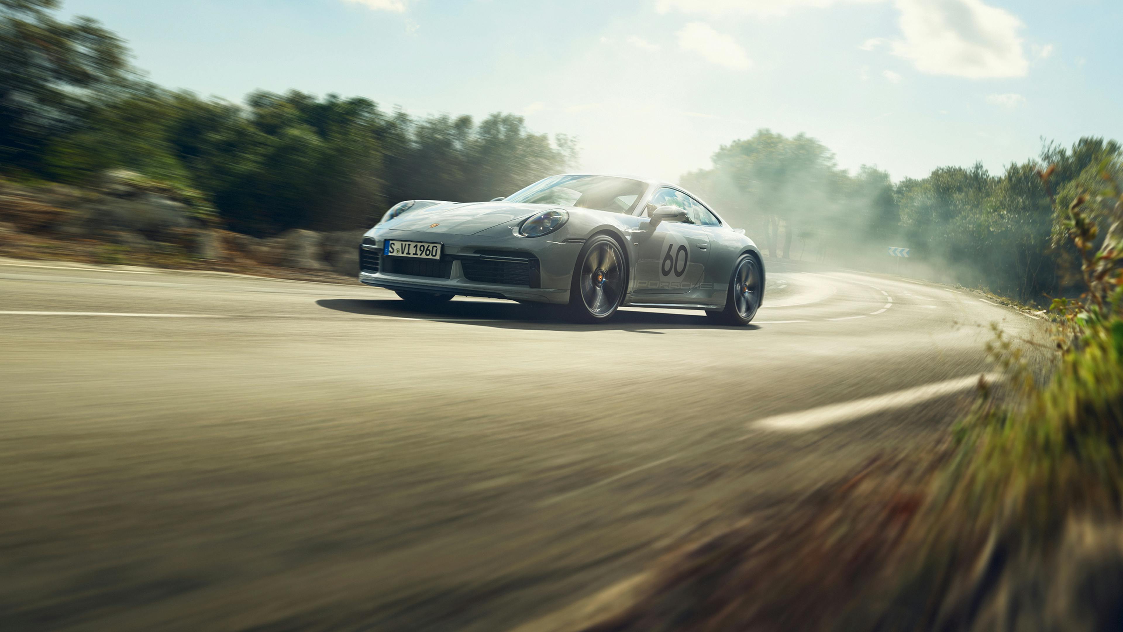 A Porsche 911 Sport Classic in Sport Grey Metallic driving along a road through the countryside. View of the front and side.