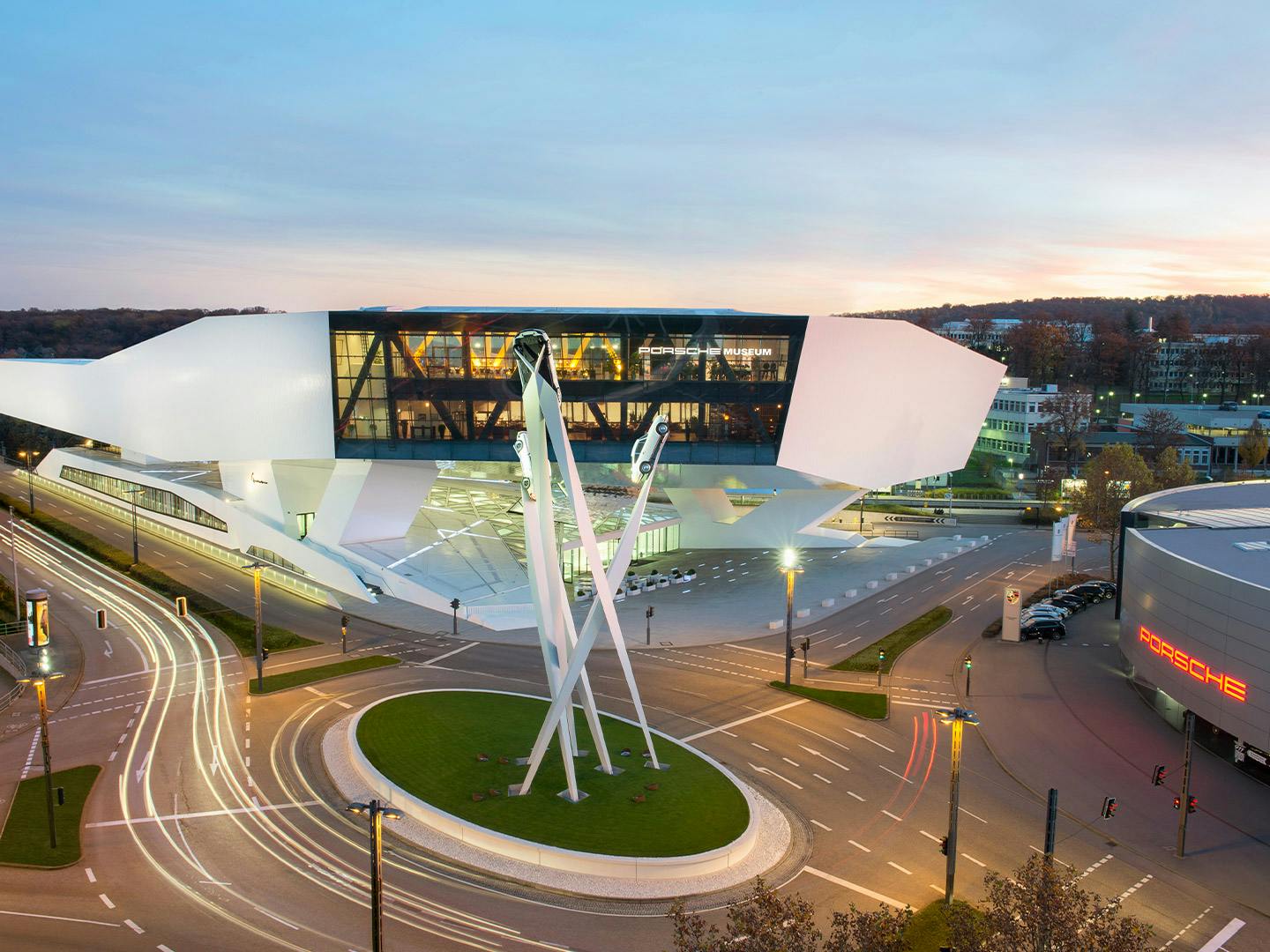 Vue aérienne d’un rond-point au centre duquel se trouve une grande sculpture de voiture, entourée de bâtiments modernes, au coucher du soleil.