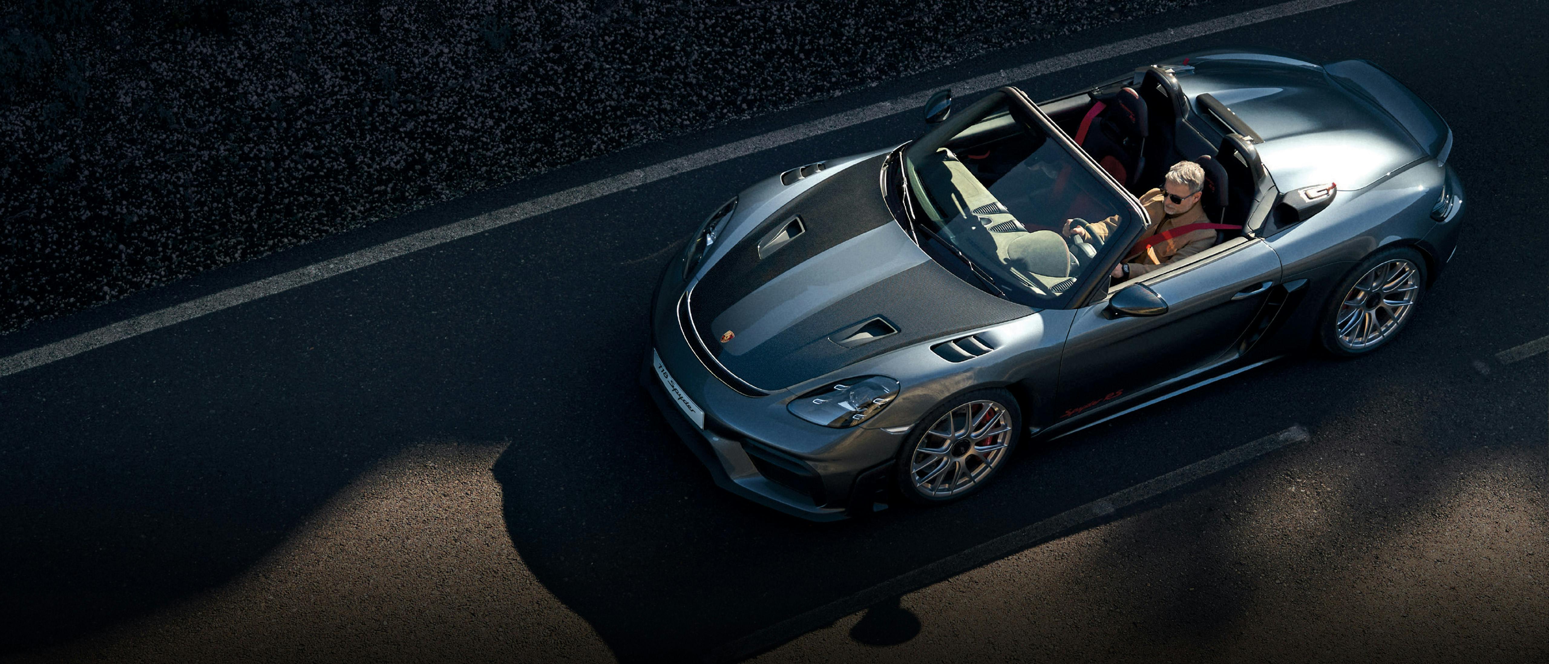 A sleek Porsche convertible in metallic gray driving on a road, showcasing aerodynamic design, red seat belts, and sporty features under sunlight.