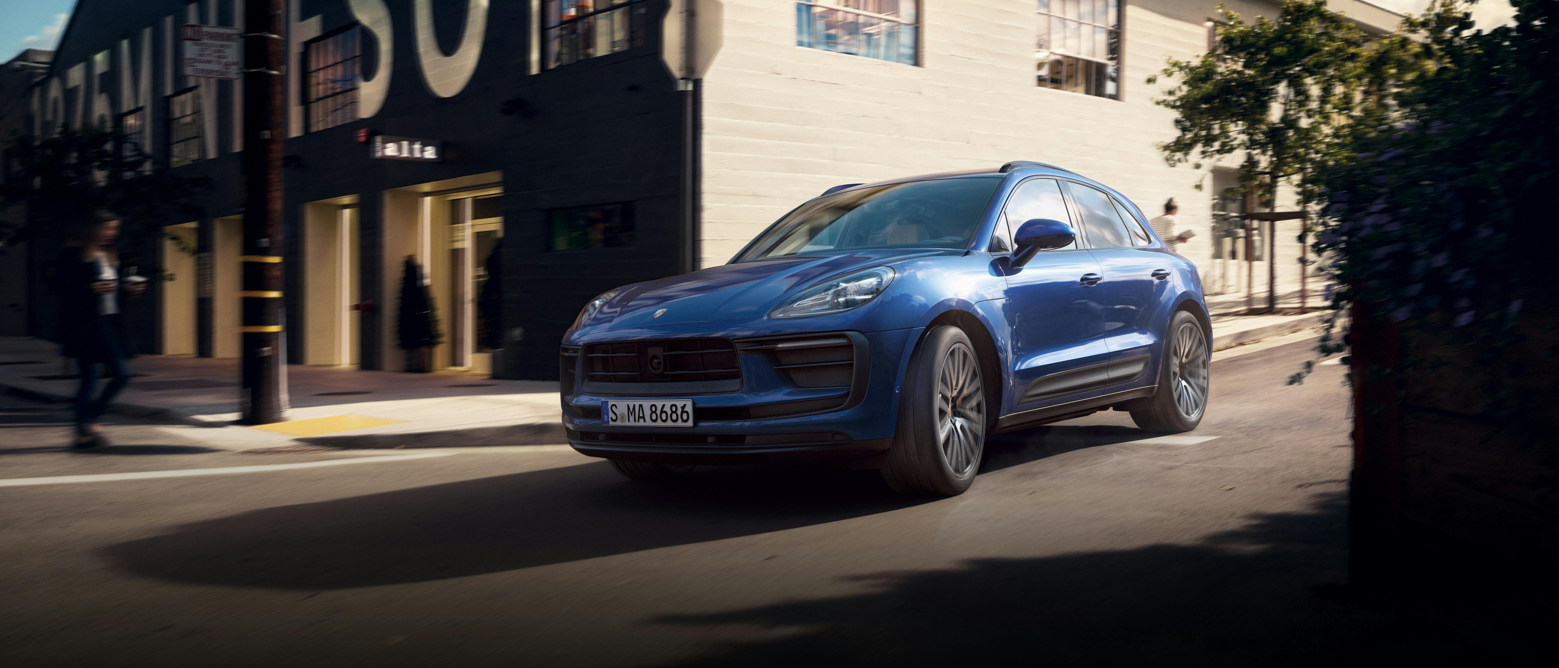 The Macan driving along a road, going past a building that a white wall on one side and a brown wall on the front.