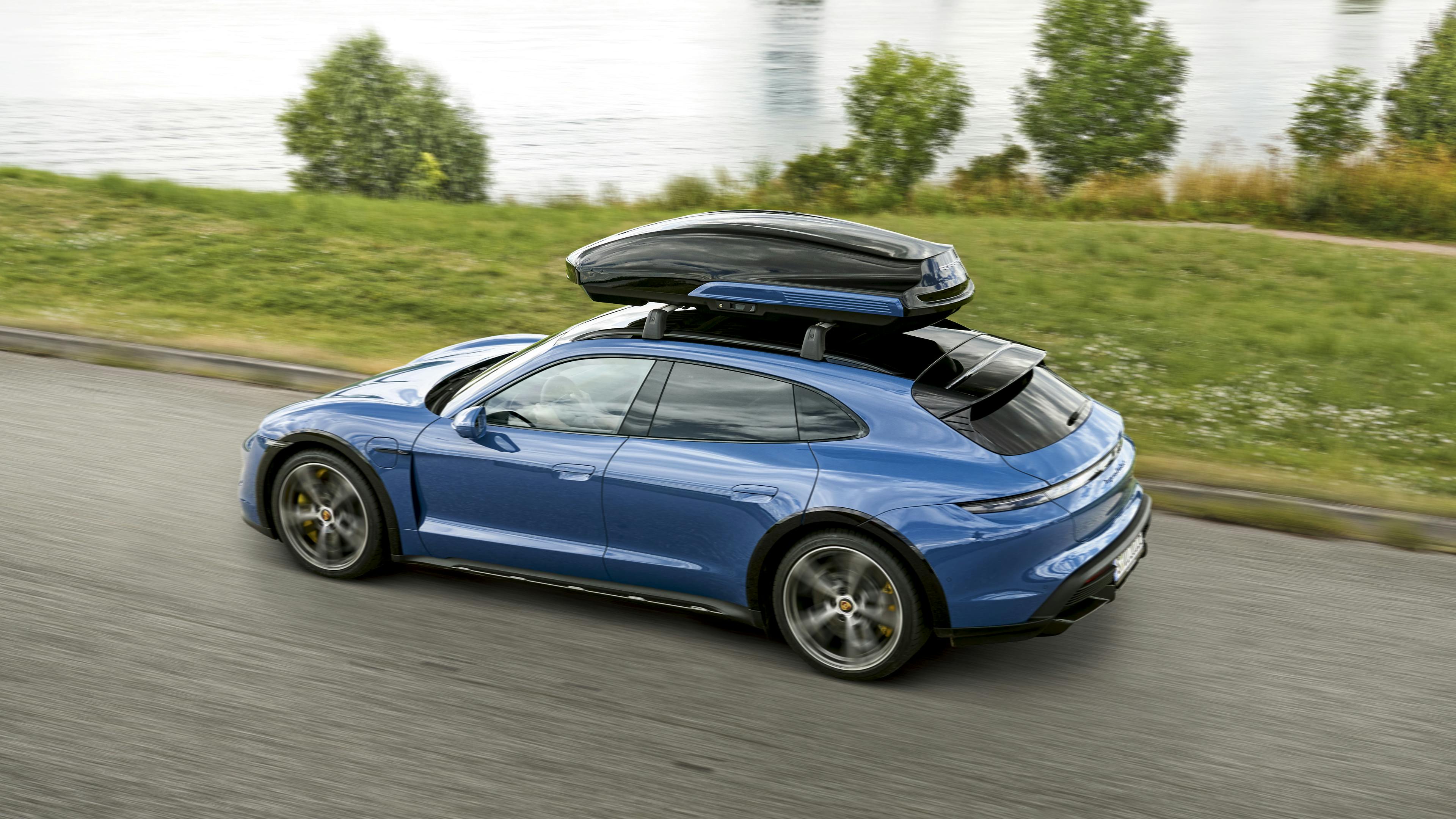 Porsche Taycan Turbo S Cross Turismo en Bleu Neptune, roulant le long d’un lac, équipée d’un coffre de toit Porsche Performance.