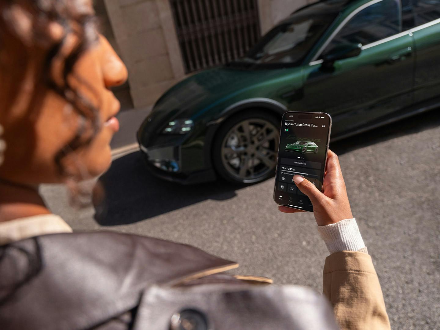 Woman holding a smartphone with the Porsche app in front of a dark green Porsche Taycan Turbo Cross Turismo on a sunny city street.