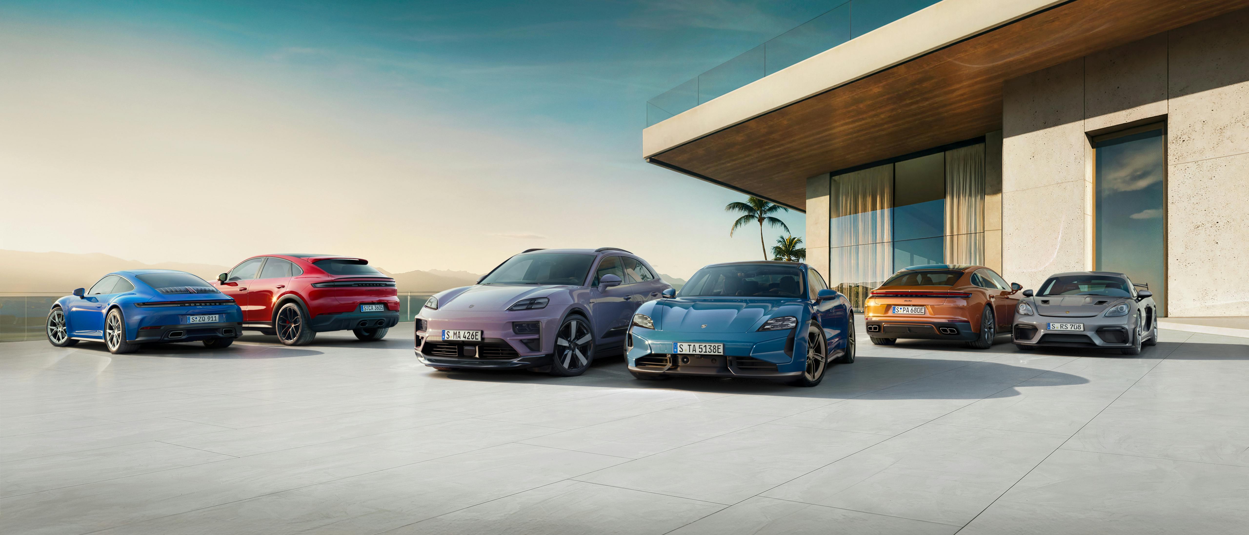 Several Porsche models, including modern electric and sport variants, parked in front of a contemporary glass-fronted building with palm trees and a warm sunset sky in the background.