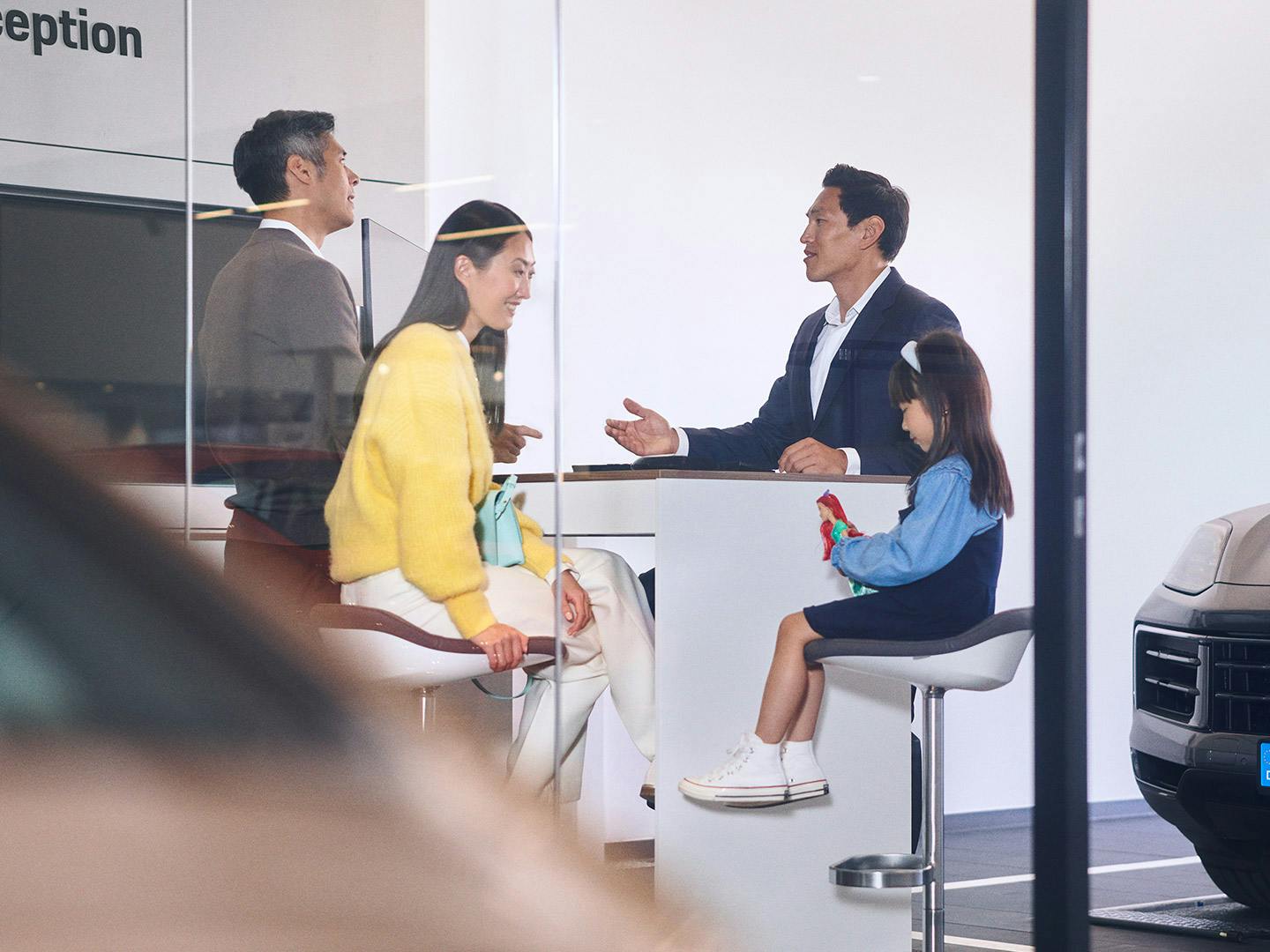 At the reception of a Porsche service centre, a customer is talking to a service advisor. A woman walks past in the background.