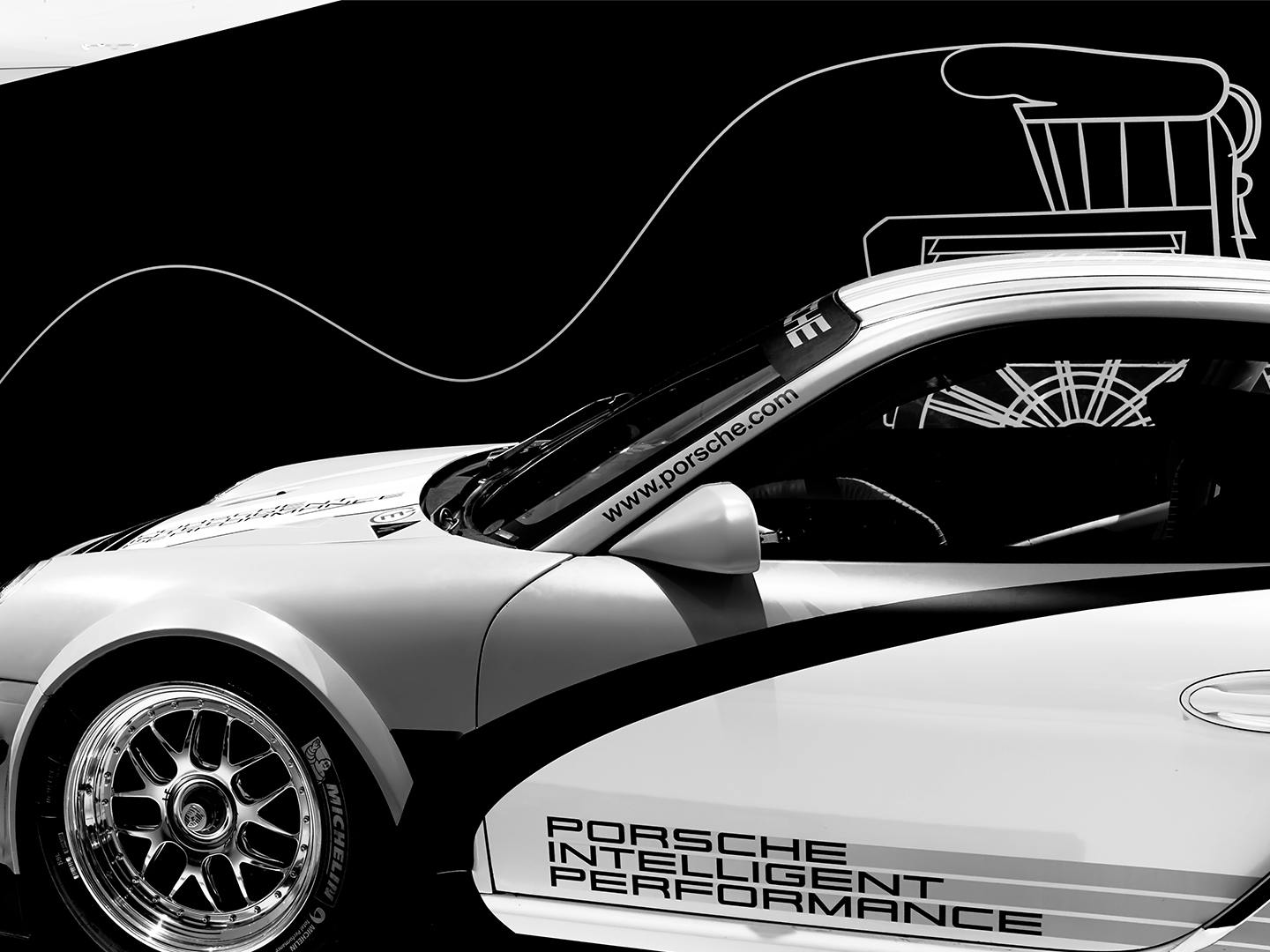 Teilansicht eines Porsche‑Rennwagens vor schwarzer Ausstellungswand mit grafischen Linien und technischen Illustrationen im Hintergrund.