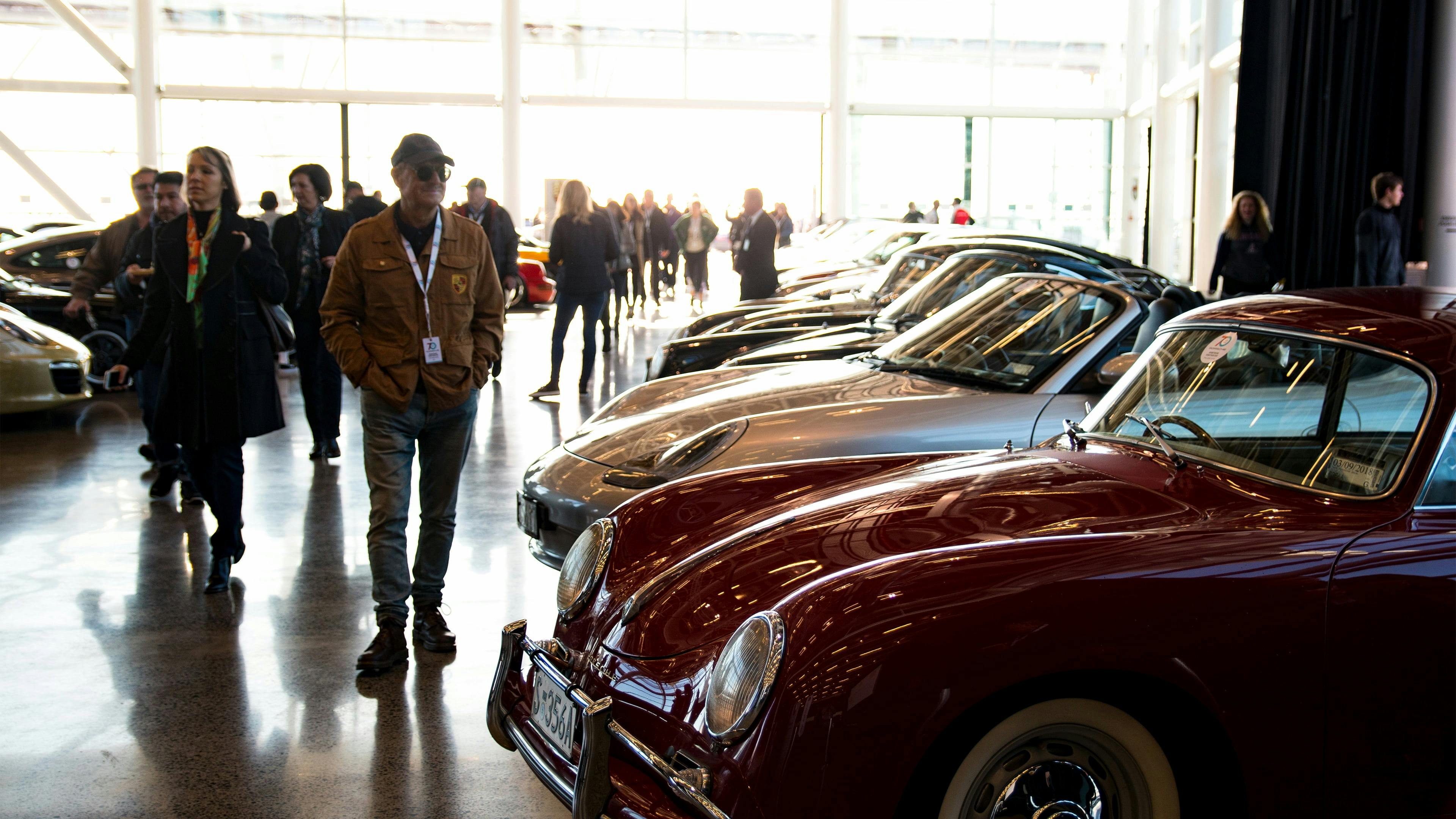 White Porsche parked on a waterfront beside a modern venue, with sports cars and event flags in the background.